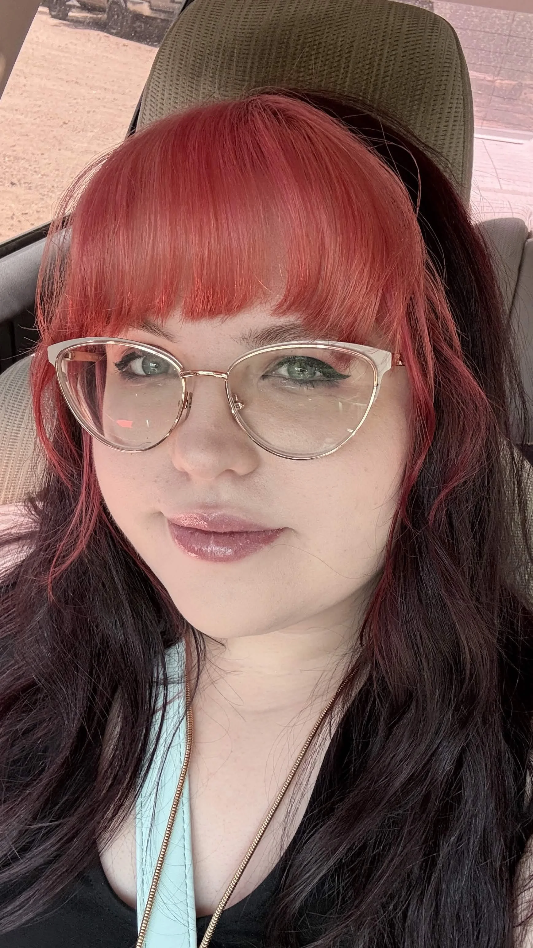 A woman with long dark hair with red highlights, wearing glasses and a black top, sitting in a vehicle. She has makeup with winged eyeliner and glossy lips, and is smiling slightly.