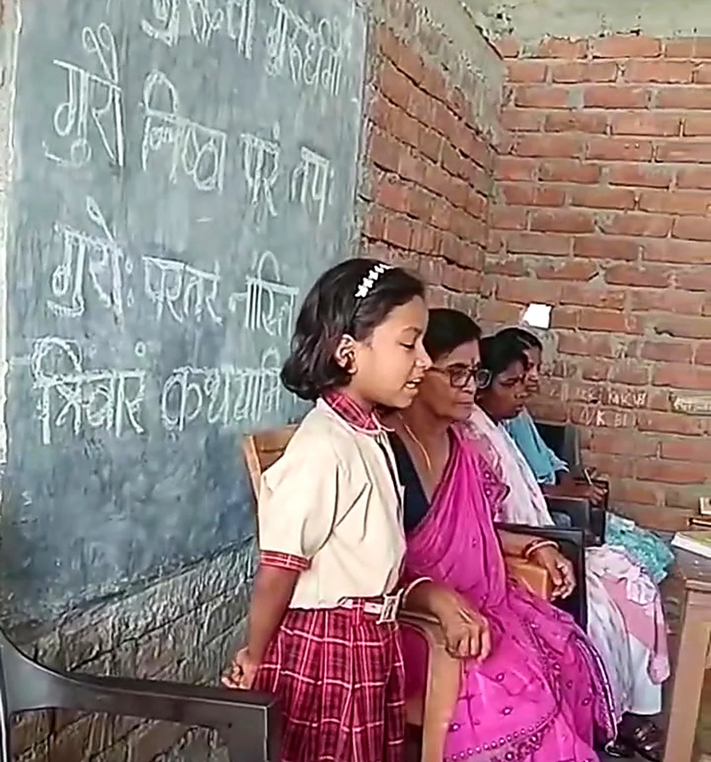 A young girl and three women sitting on chairs against a brick wall and a chalkboard with writing in Hindi. The girl is wearing a school uniform and the women are dressed in traditional Indian clothing.