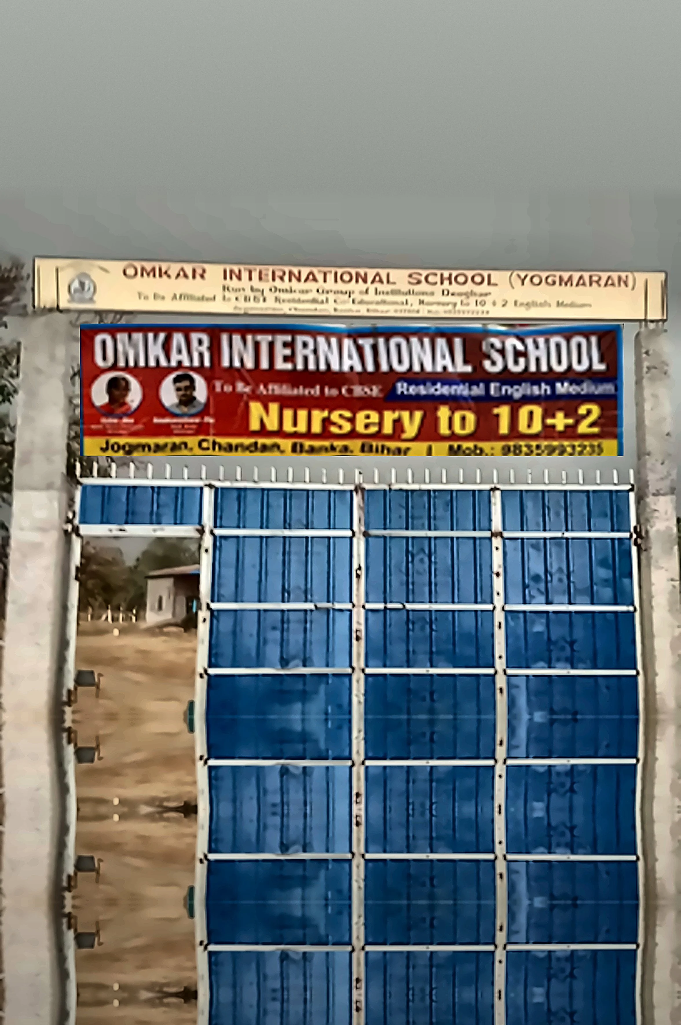 Entrance gate of Omkar International School, with a signboard displaying school name and details, metal gate painted blue, with a view of a dirt road and some buildings in the background.