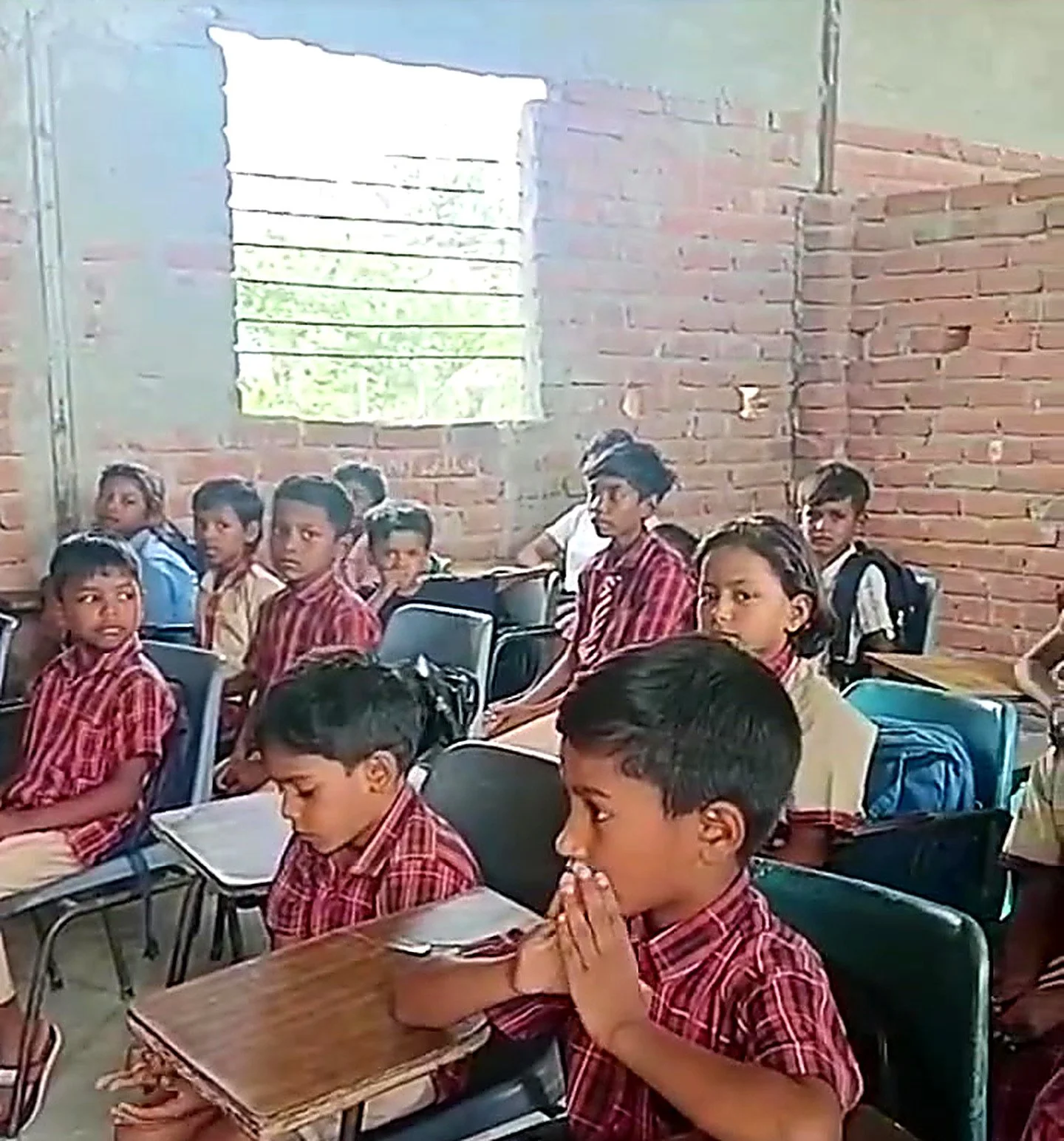 A classroom full of young students sitting at desks, some with hands in a prayer position and others looking towards the front, with an unfinished brick wall and a window in the background.