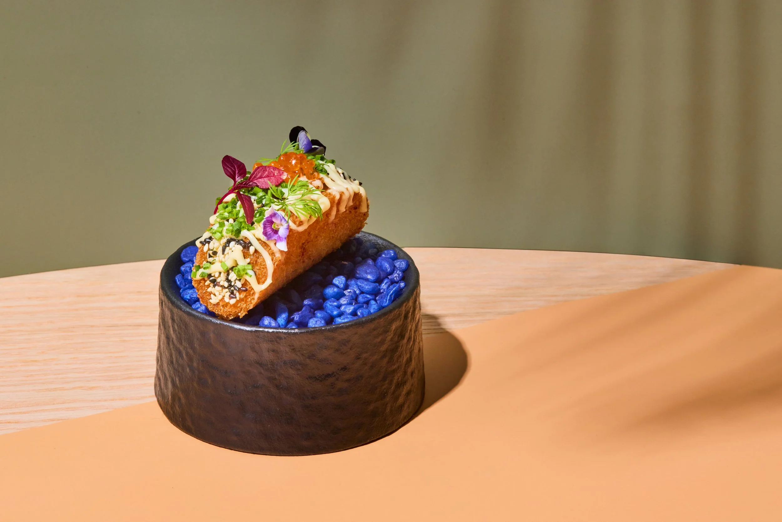 A plated dessert featuring a cannoli pastry topped with colorful edible flowers, green herbs, orange fish roe, black and white sesame seeds, and a drizzle of white sauce, served on a black textured bowl filled with blue decorative stones on a light wooden surface.