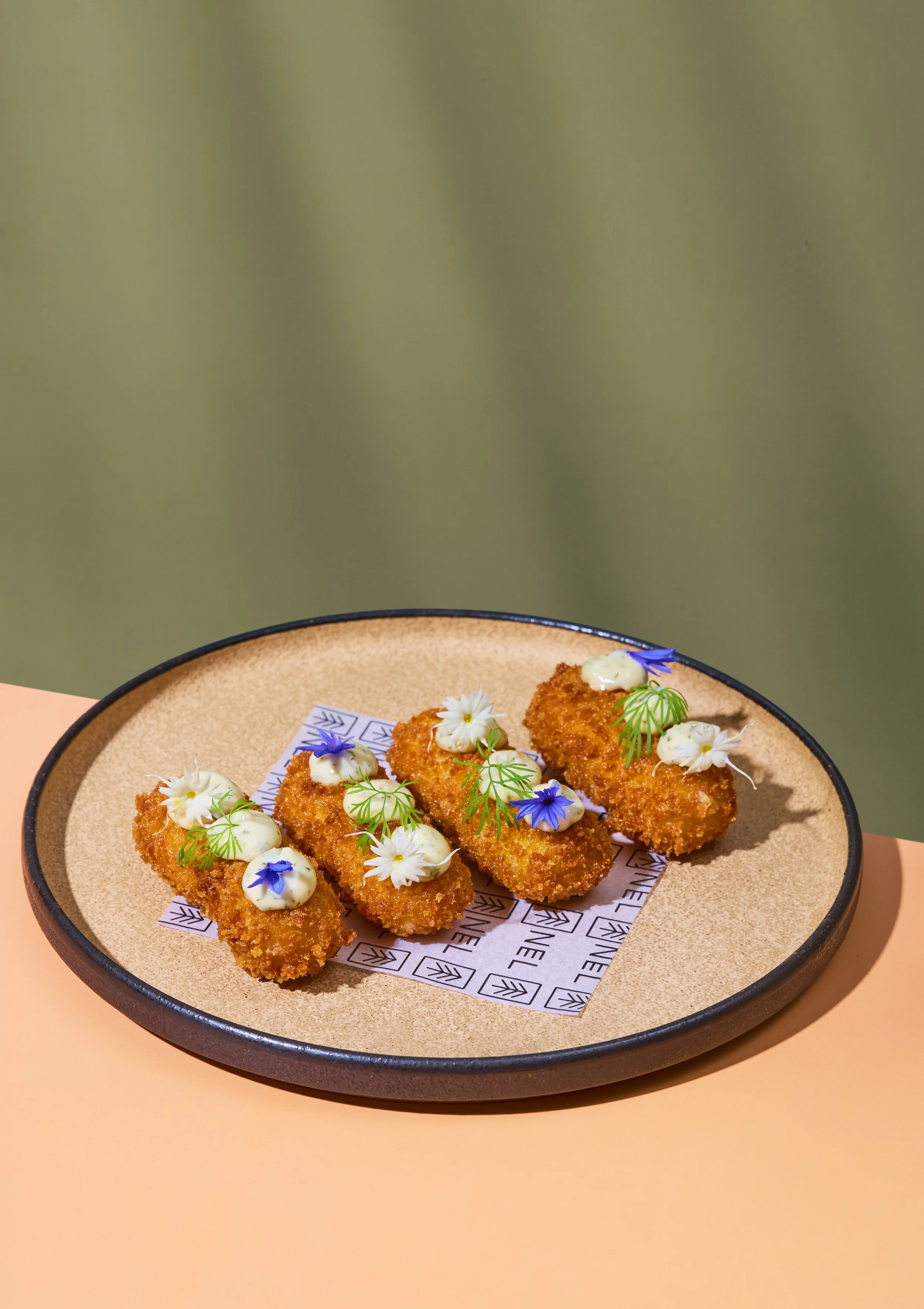 Fried food bites garnished with edible flowers and herbs, served on a round ceramic plate with branded paper lining.