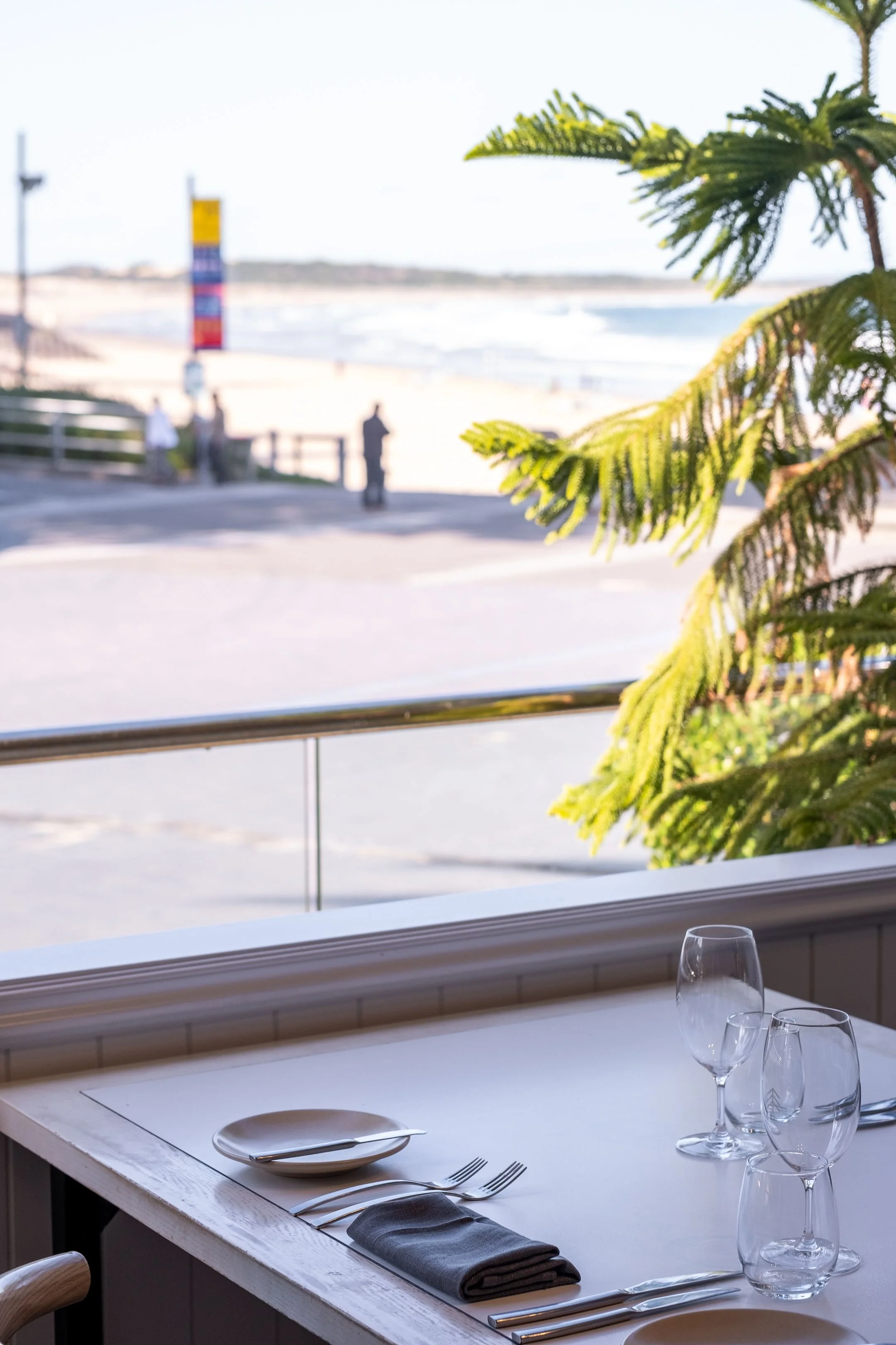 Table set for dining with plates, forks, napkins, and wine glasses, overlooking a beach with ocean, sand, and palm trees in the distance.