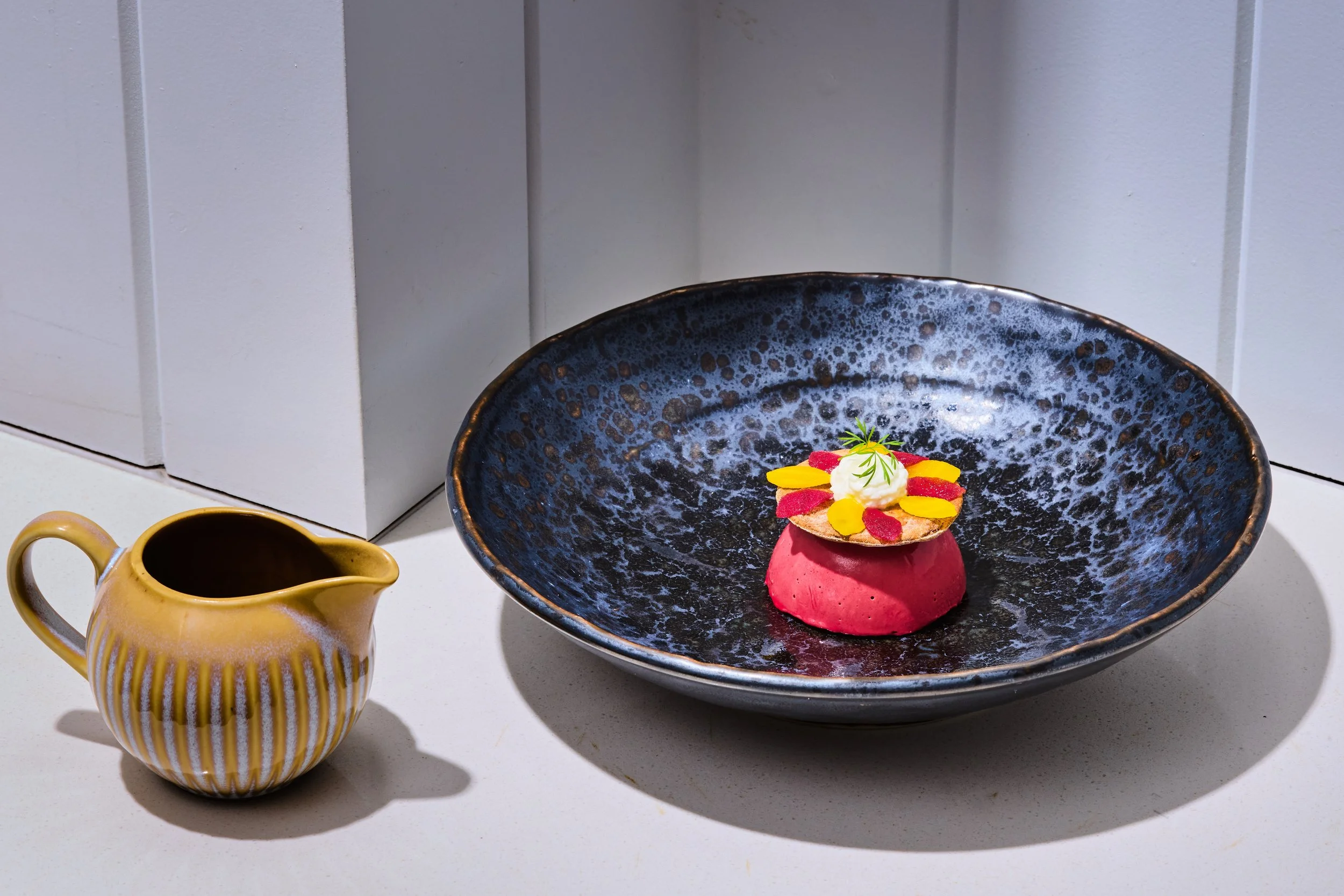A yellow and white striped ceramic pitcher next to a black textured bowl containing a small pink dessert topped with whipped cream, yellow petals, red petals, and a green herb, with a white cabinet in the background.