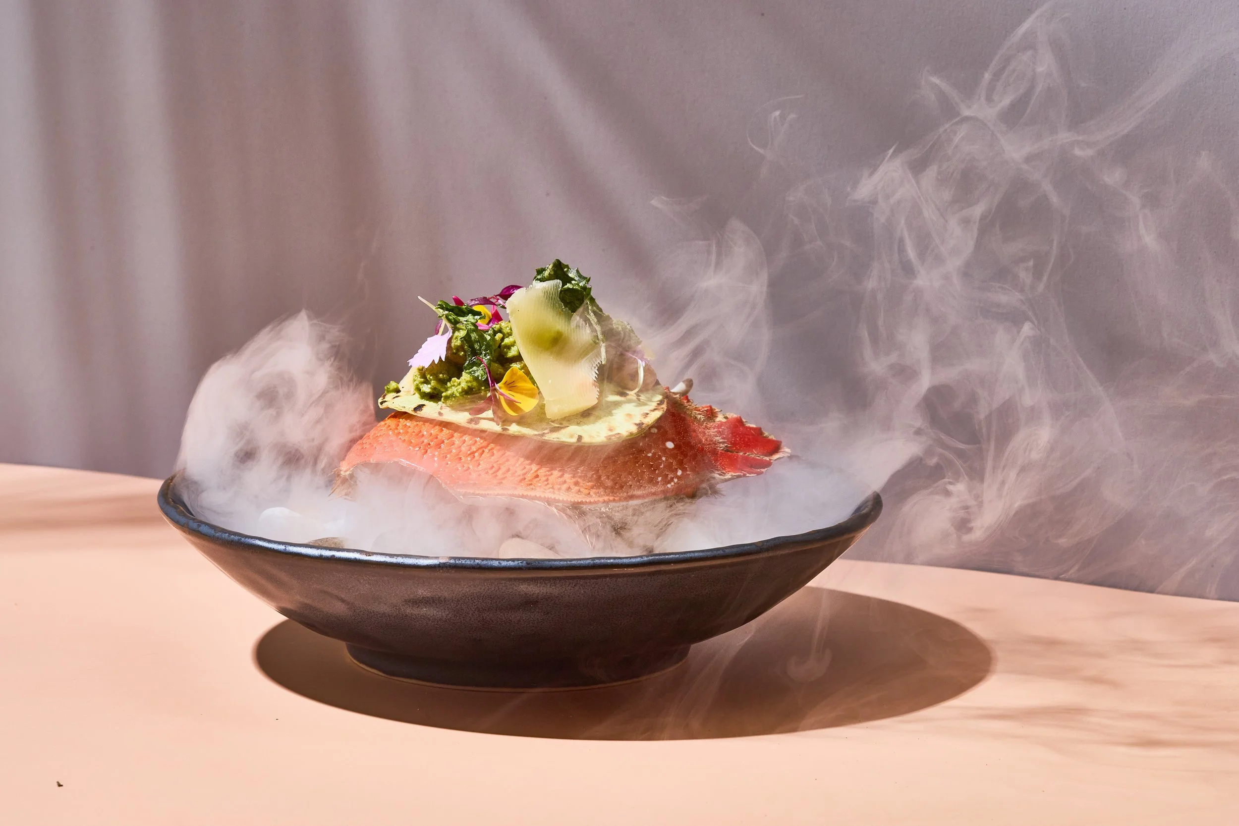 A plate with a fillet of fish topped with herbs, cheese, and edible flowers, surrounded by aromatic smoke.