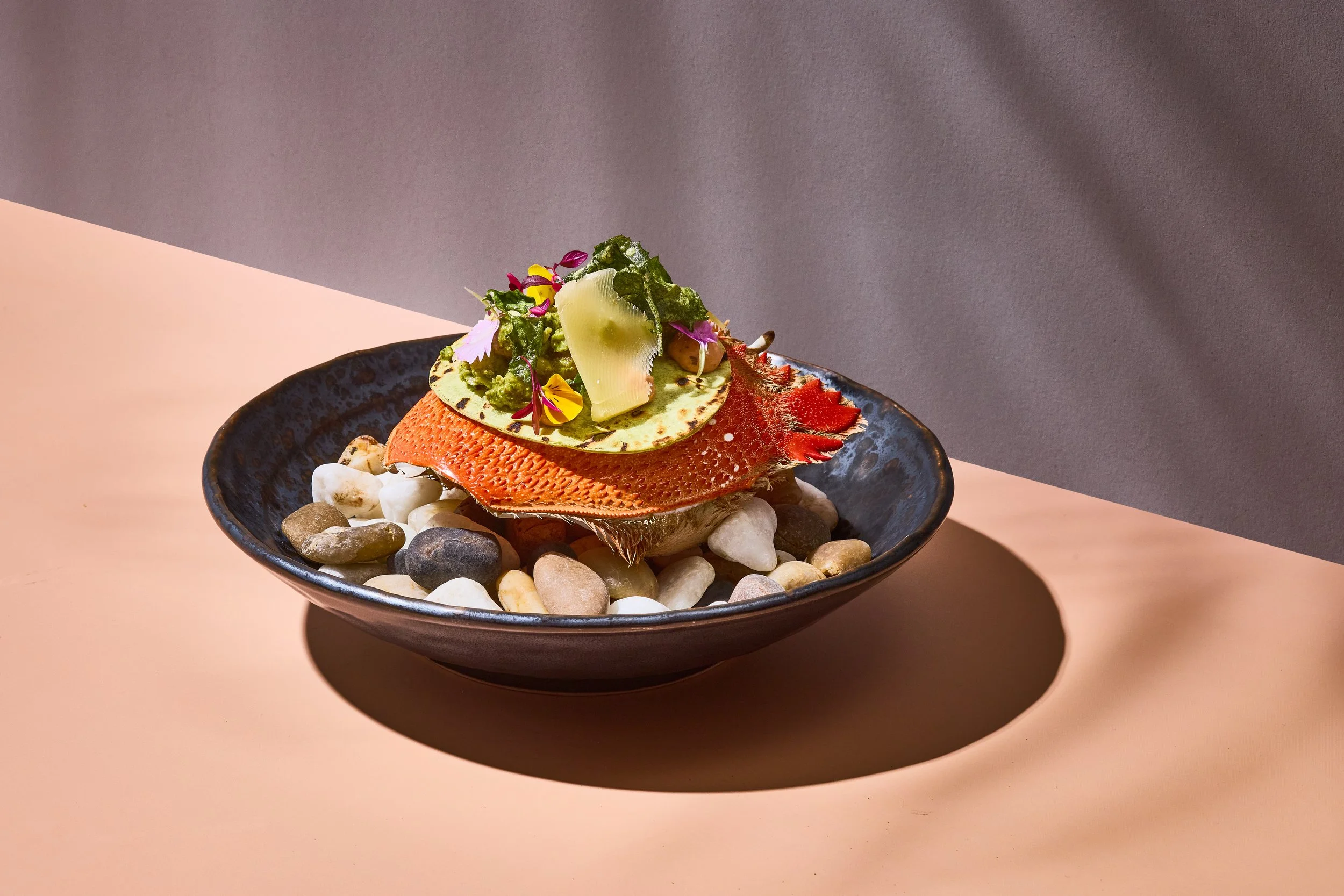 A black bowl with a textured surface, filled with a seafood dish including a crab shell and garnished with edible flowers, herbs, and lemon slices, placed on a bed of mixed smooth pebbles.