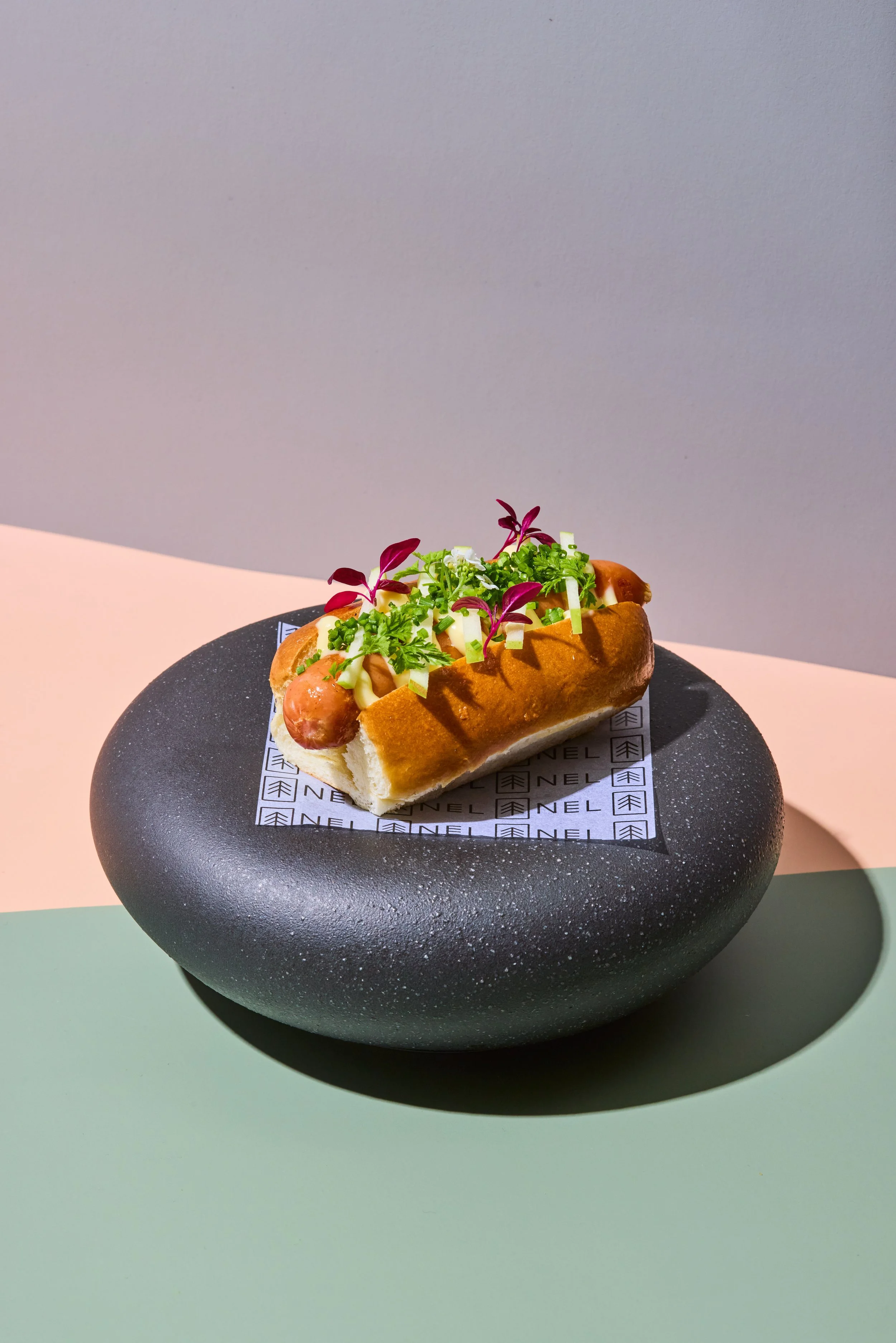 A hot dog in a bun topped with chopped green onions, microgreens, and small purple leaves, served on a black plate with branded paper.