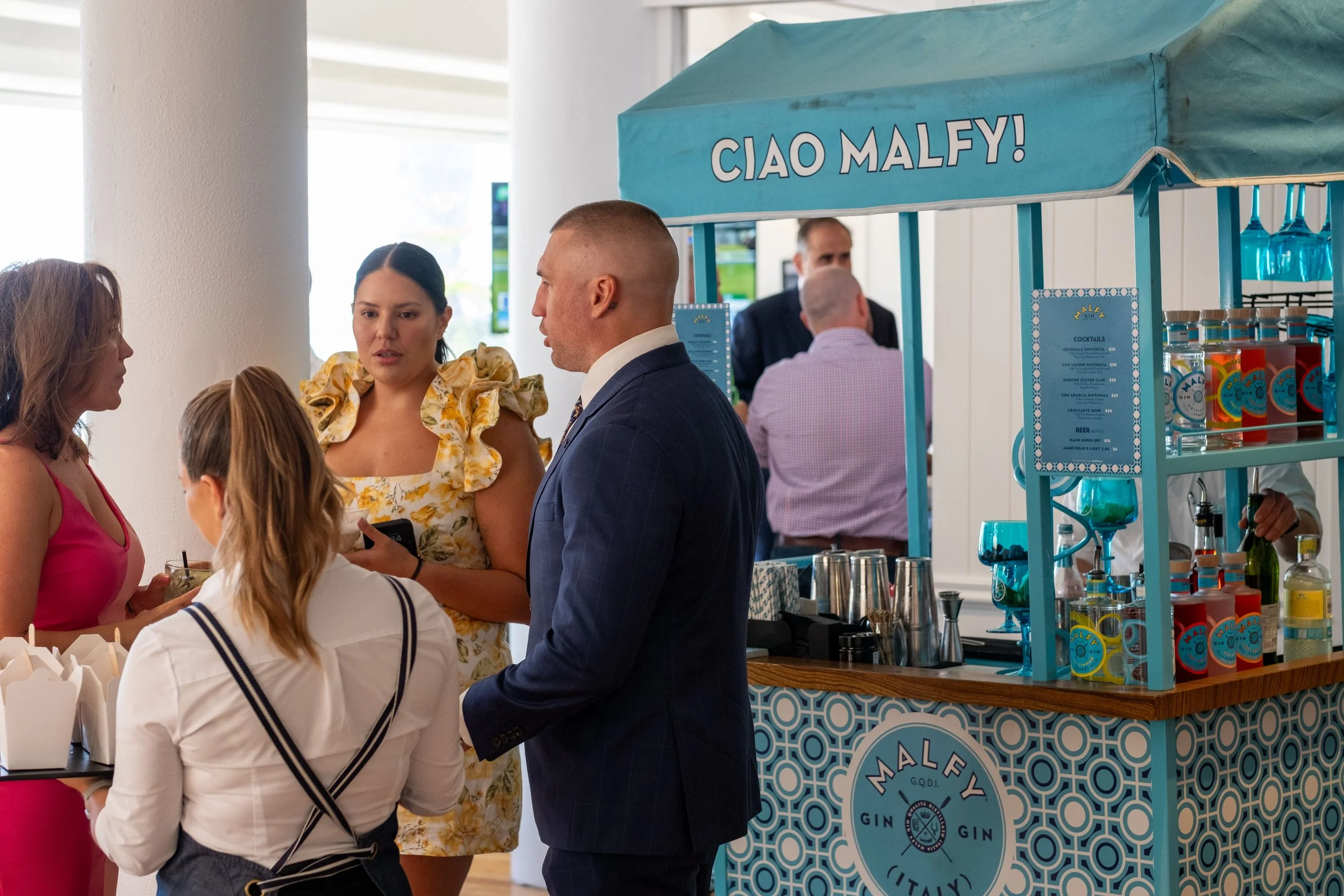 People gathered around a blue Malfy Gin stand at an event, engaging in conversation, with a woman wearing a pink dress, a woman in a white striped shirt, and a man in a blue suit visible. The stand displays bottles of Malfy Gin and a menu.