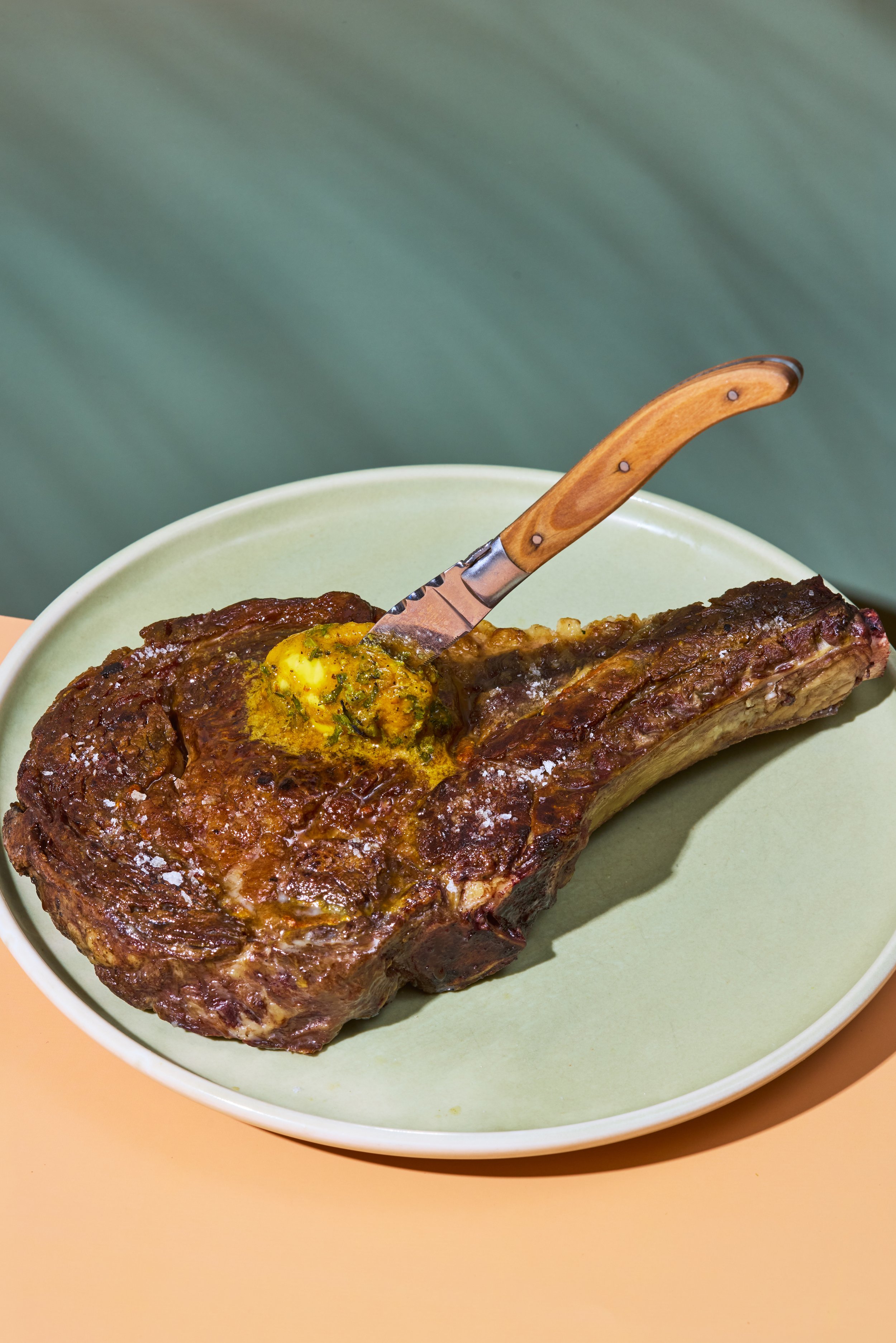 A large, cooked beef ribeye steak on a green bowl, topped with herb butter and a carving fork with a wooden handle.