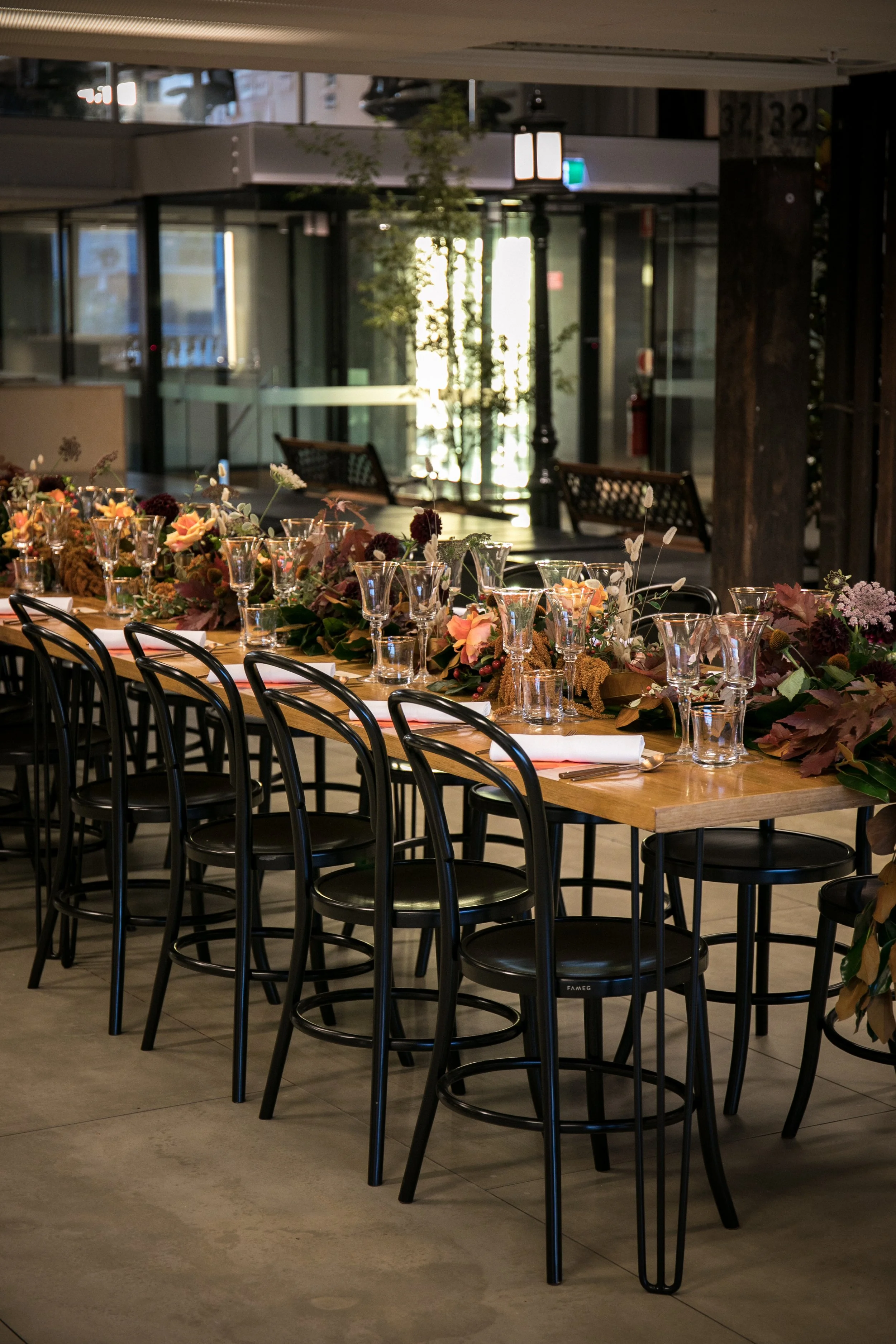 A long dining table decorated with flowers and glassware in a modern restaurant setting with large windows and outdoor views.