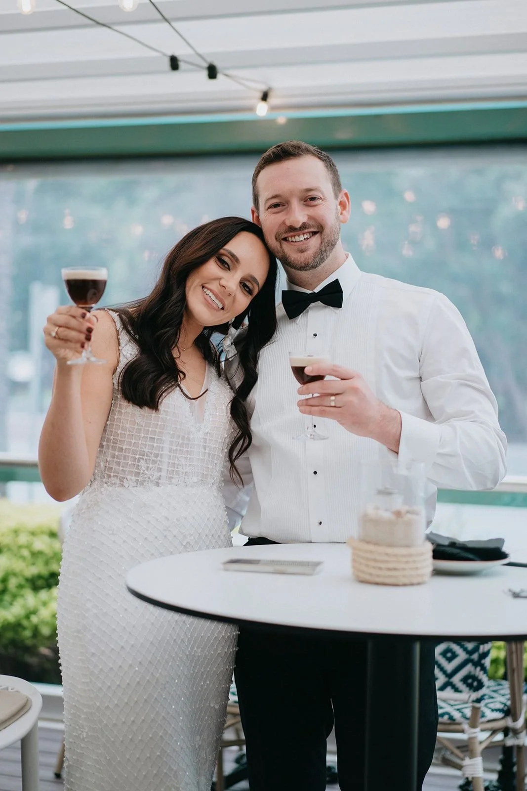 A newlywed couple dressed in wedding attire, smiling and holding glasses of red wine, standing close together at a celebration event.