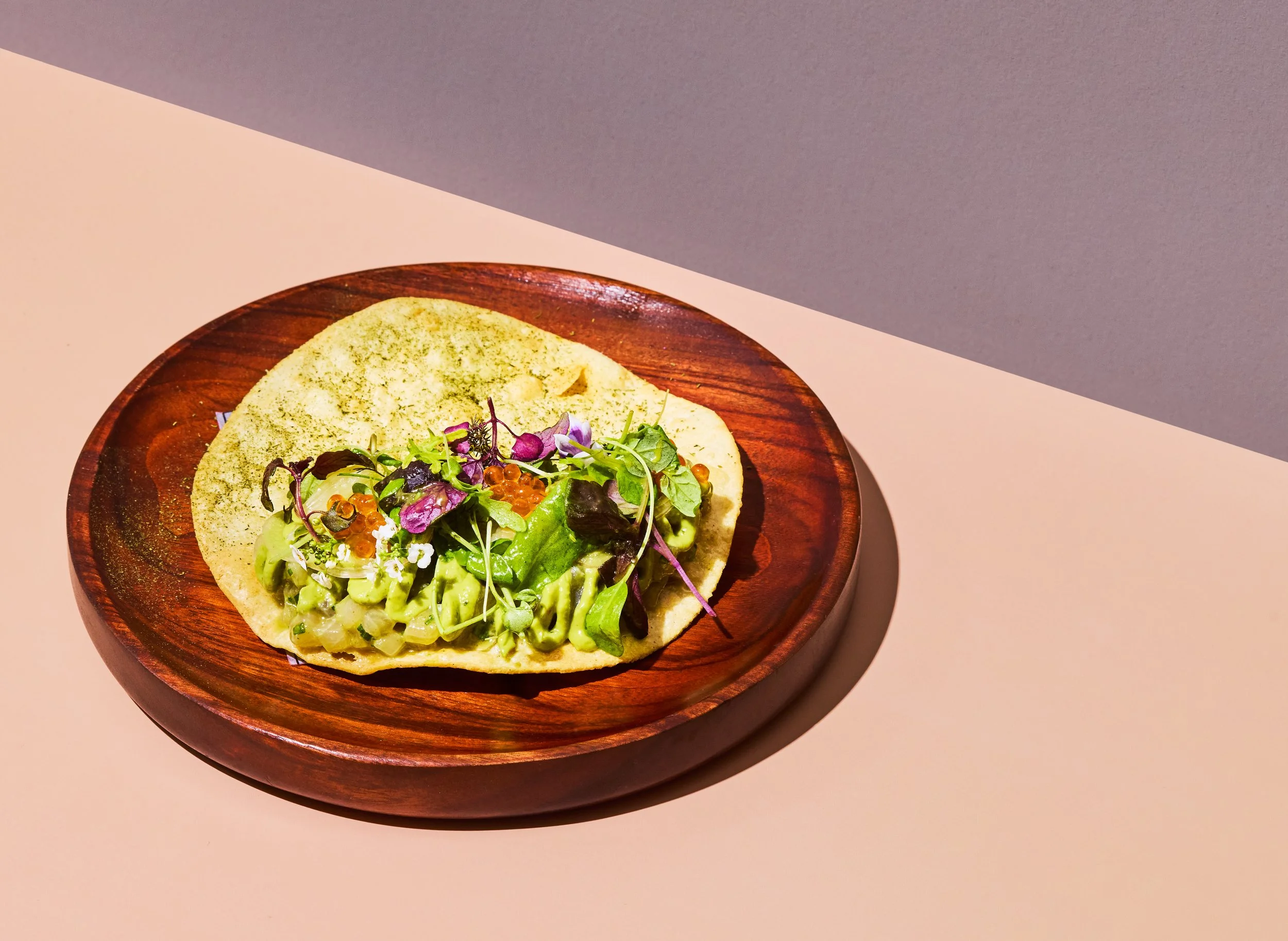 A wooden plate with a flatbread topped with avocado, mixed greens, microgreens, and red caviar, on a two-tone background.