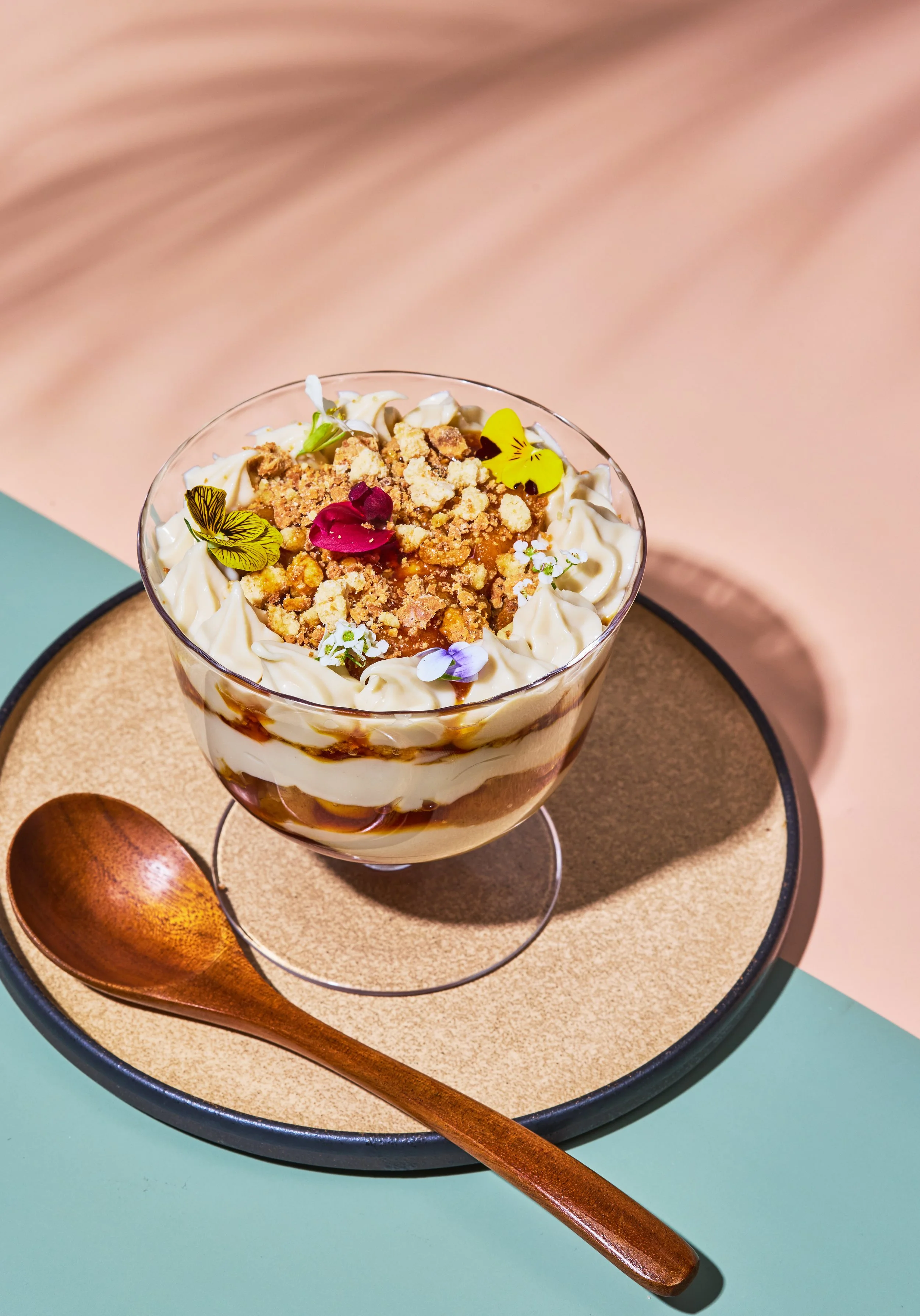 Close-up of a dessert in a glass, topped with crumbles, edible flowers, and decorative yellow butterflies. The dessert sits on a round tray with a wooden spoon.