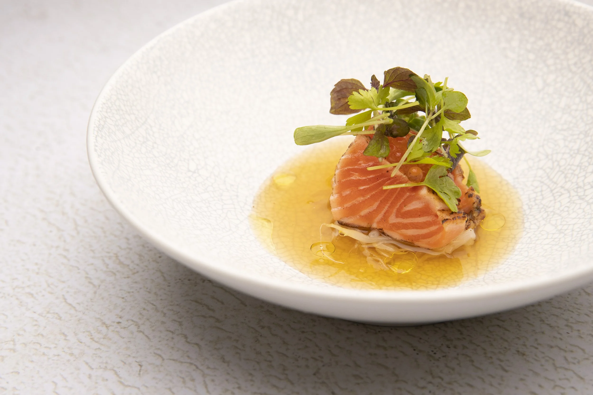 A plated dish featuring a piece of cooked salmon garnished with microgreens and drizzled with a light sauce, served on a white textured plate.