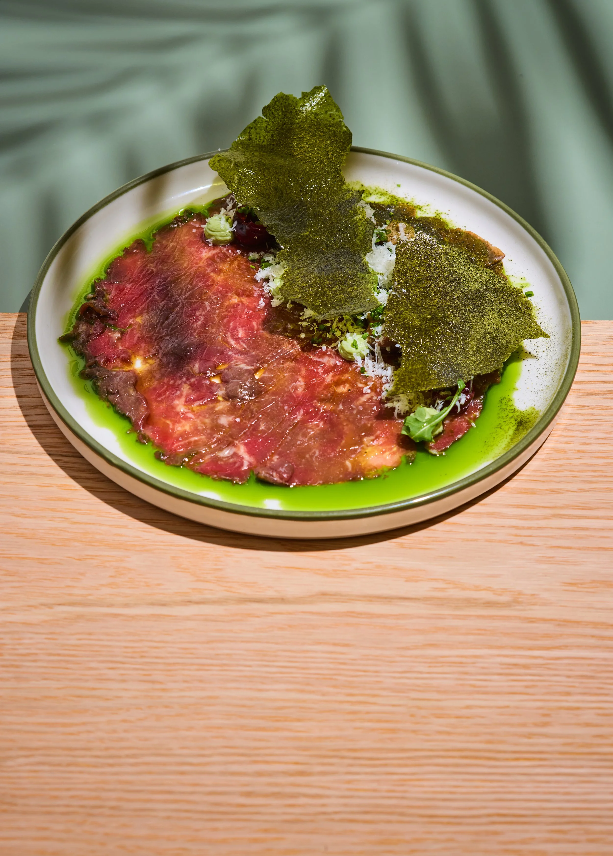 Thin slices of raw beef carpaccio garnished with herbs, microgreens, green sauce, and crispy seaweed on a white plate with a green sauce drizzle around the edge.