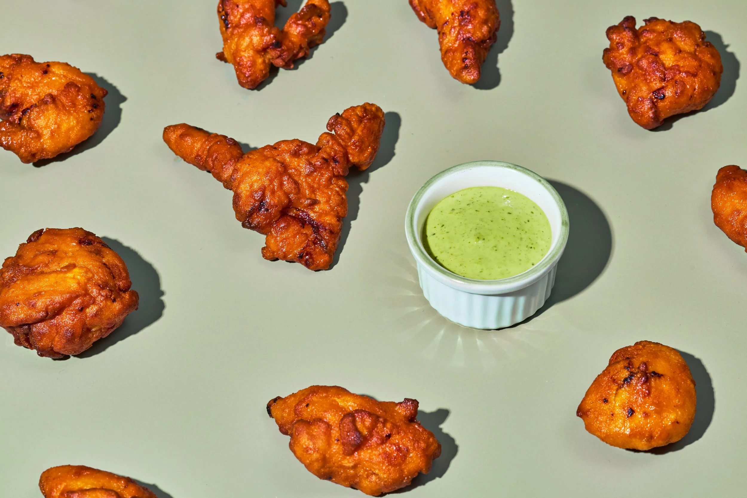 Fried chicken pieces with a side of green dipping sauce in a small white bowl on a light-colored surface.