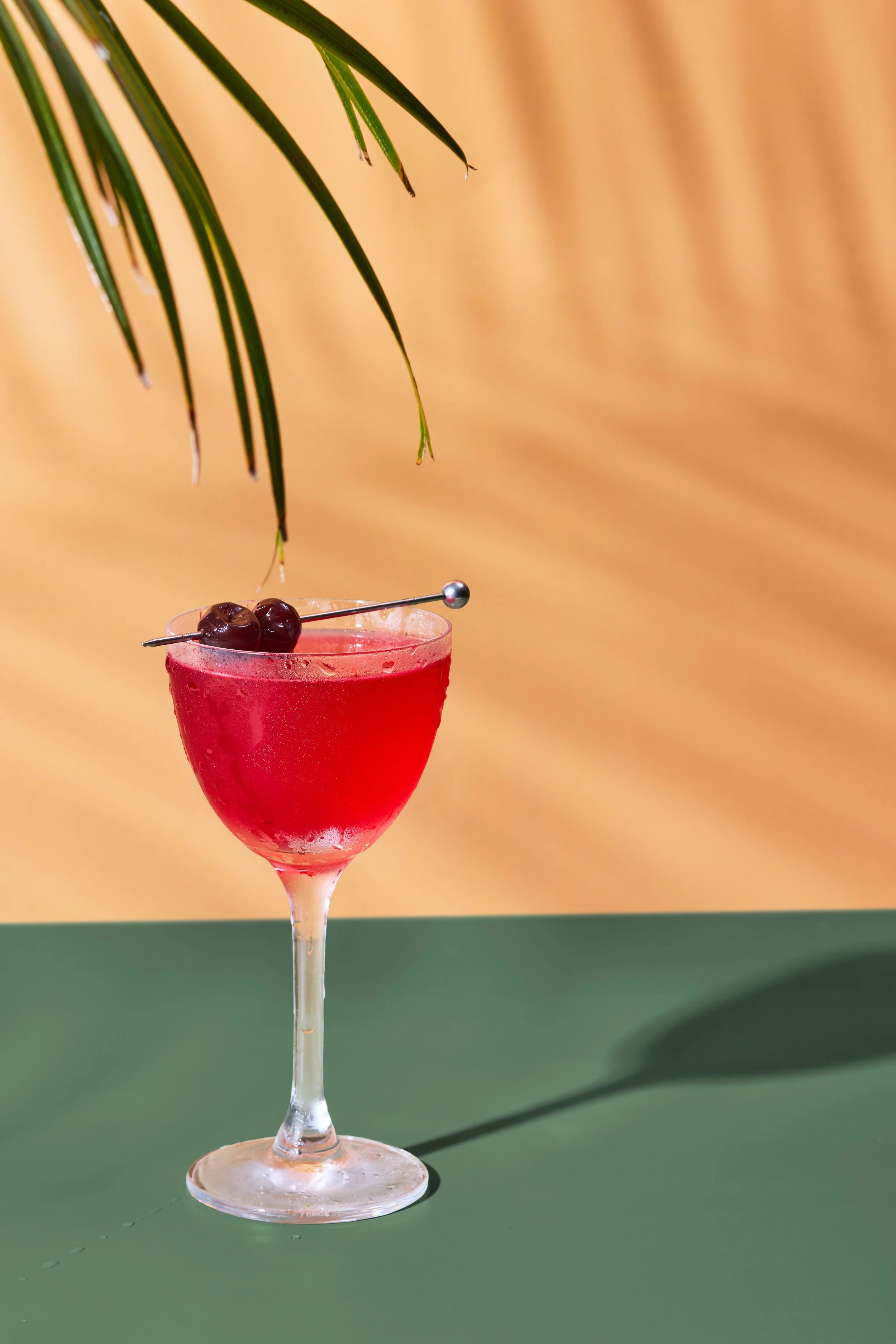 A red cocktail glass with a cherry garnish on a toothpick, set against a background with a yellow upper section and green lower section, with a green leafy branch hanging overhead.