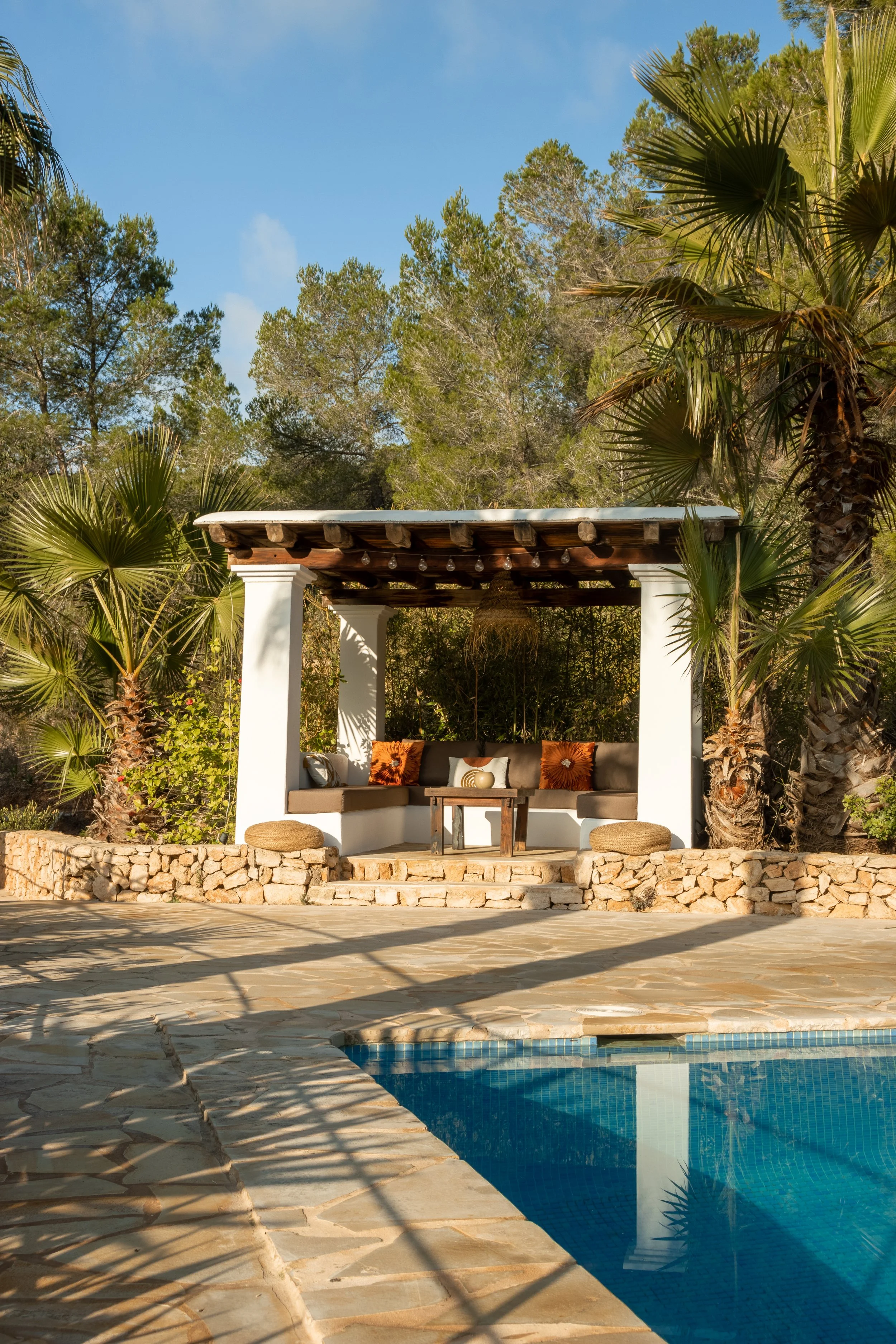 Outdoor pool with stone deck featuring a shaded seating area with cushioned benches, pillows, and a small table, surrounded by palm trees and lush greenery under a blue sky.