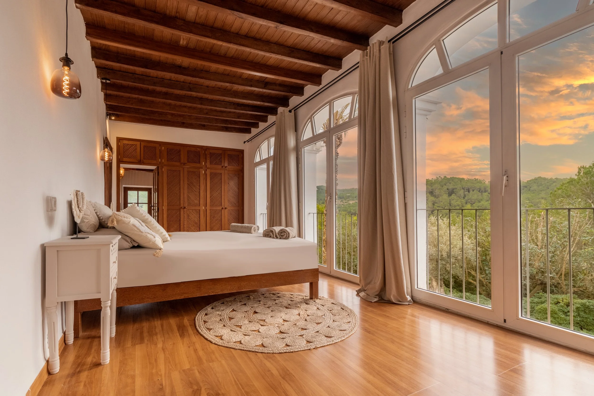 A bedroom with a large bed, wooden ceiling beams, white walls, polished wooden floors, a small white bedside table, and floor-to-ceiling windows with curtains showing an outdoor view of green trees and a sunset sky.