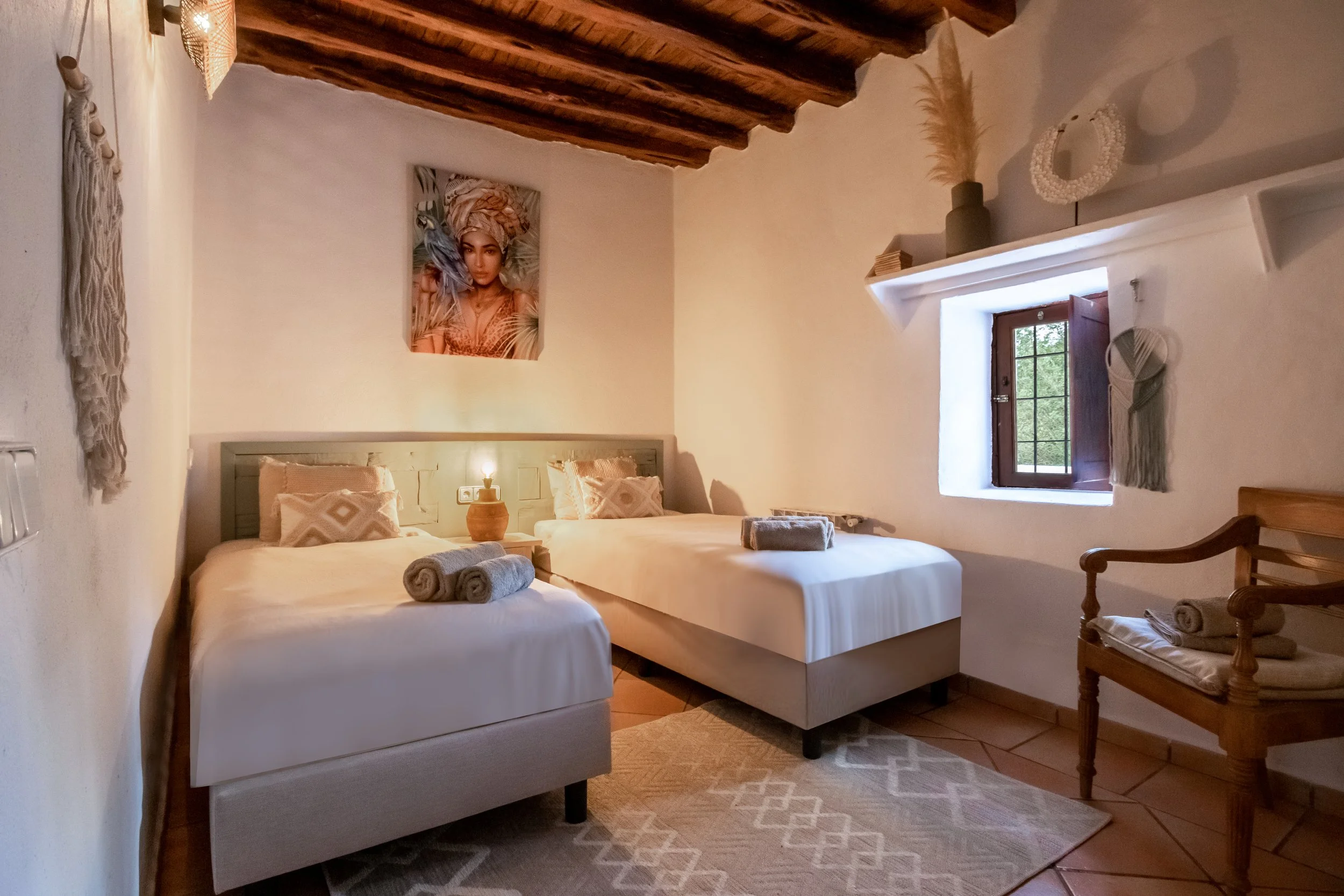 A cozy bedroom with two twin beds with white bedding, two rolled towels on each bed, a wooden nightstand with a small lamp, a framed portrait of a woman with a bird above the beds, a small window with wooden shutters, and decorative items including vases and wall hangings on the white walls. The ceiling has exposed wooden beams.