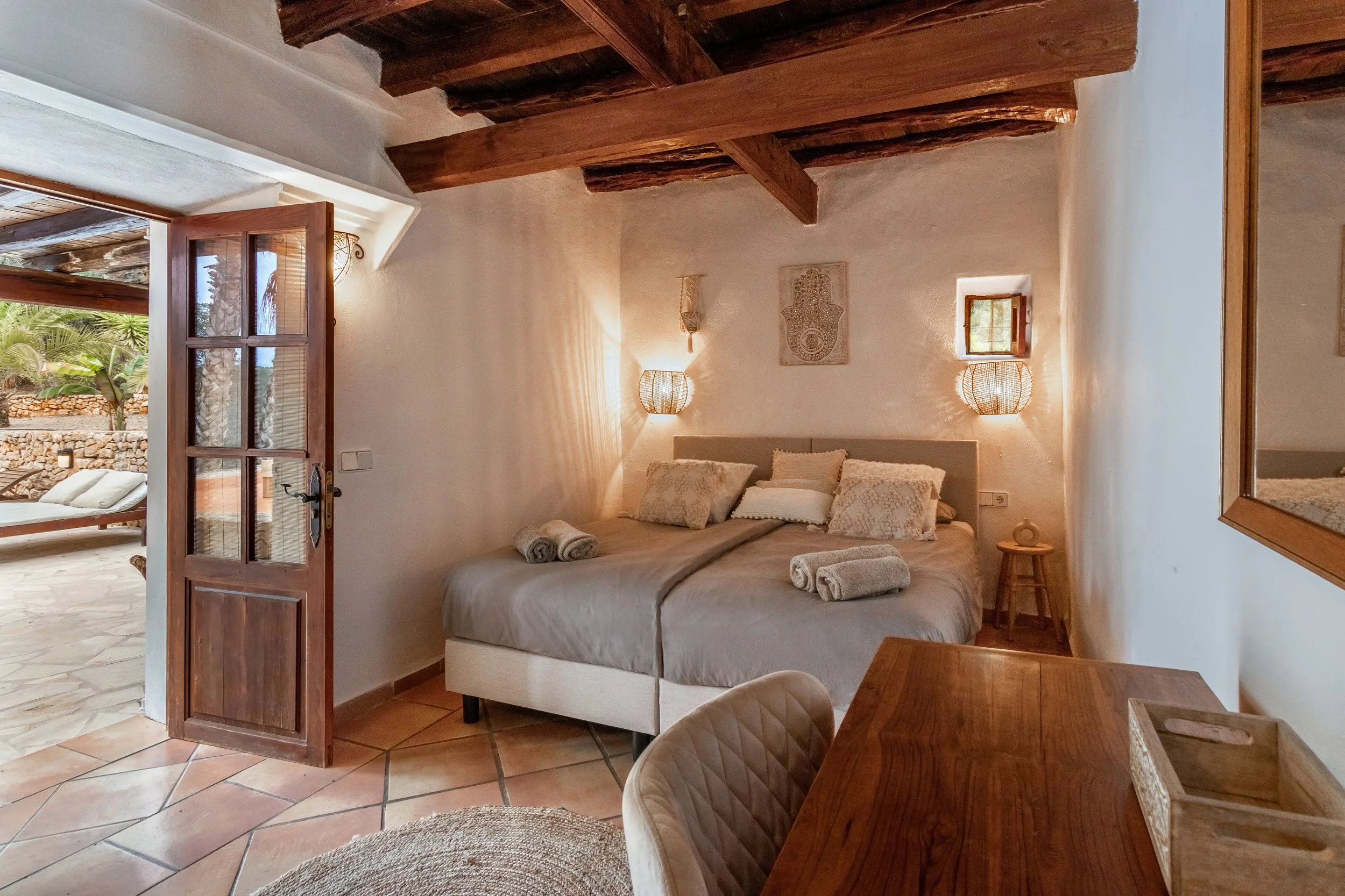 Cozy bedroom with a large bed, decorative pillows, and neatly folded towels. Warm lighting from wall lamps, wooden ceiling beams, an open door leading to an outdoor patio with lounge chairs, and a small window on the right wall.