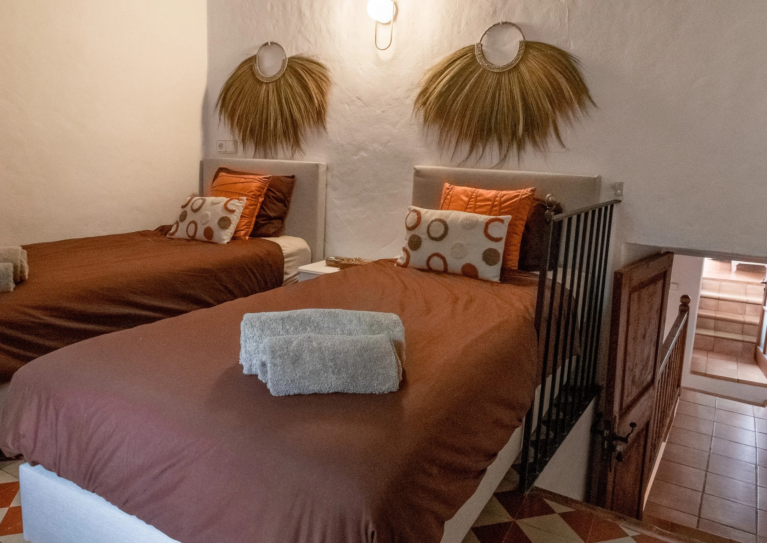 A bedroom with two beds, each with brown and orange bedding and decorative pillows. Wall-mounted hair accessories resembling large brushes with bristles are above each bed. There is a small table between the beds, a staircase leading down, and tile flooring.
