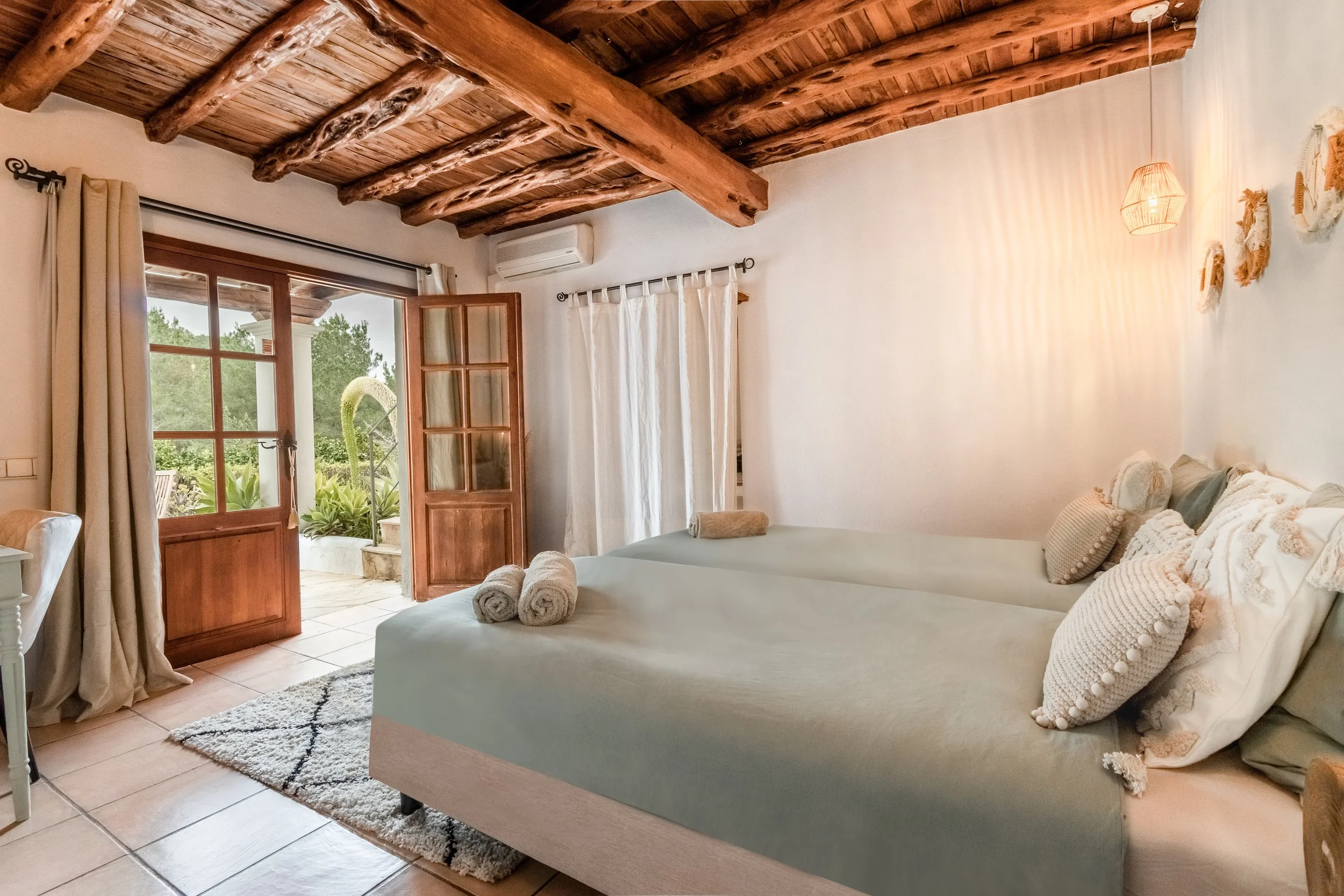 A cozy bedroom with two single beds, each with three white pillows, soft linens, and beige towels. The bedrooms opens to a patio with greenery, through wooden-framed glass doors, and has wooden ceiling beams, white walls, and curtains.