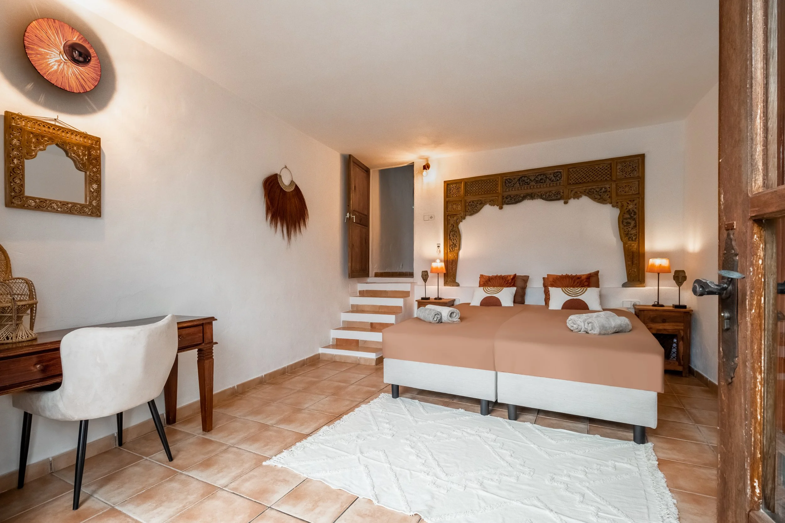 Hotel room featuring two single beds with rolled towels, wooden headboard with intricate carvings, bedside lamps, a white rug, a wooden desk with a chair, and decorative wall hangings.