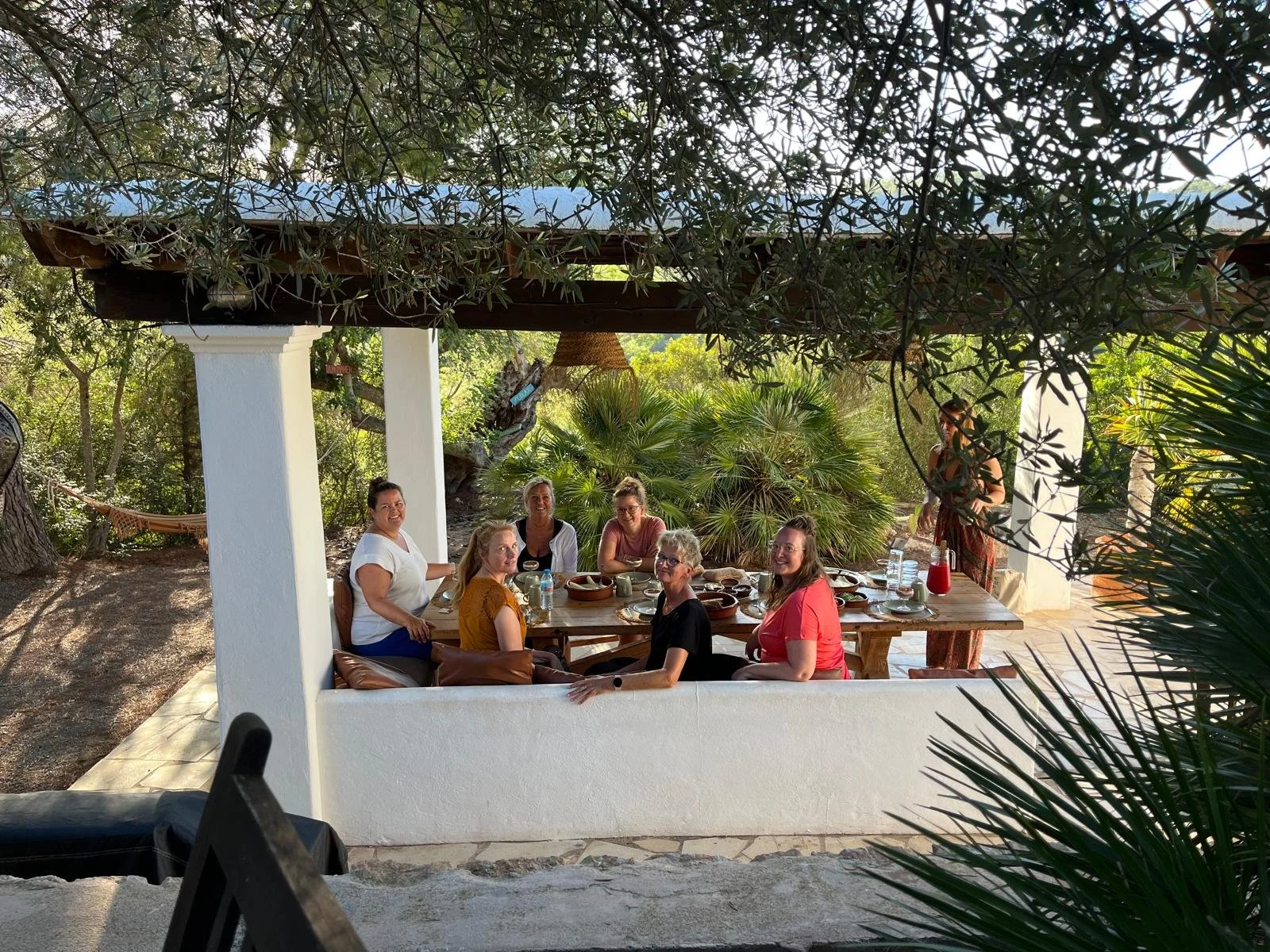 Group of women sitting around an outdoor dining table on a patio, surrounded by greenery and trees, with a woman standing nearby.