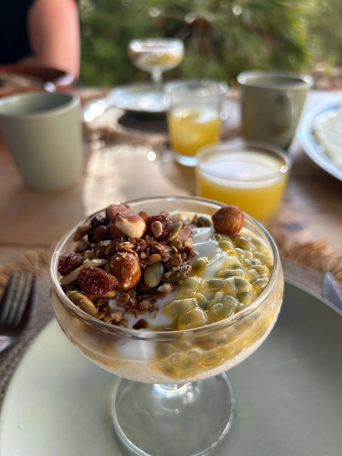 Close-up of a parfait with yogurt, granola, nuts, and seeds in a glass, on a white plate, with breakfast drinks and cups on a wooden table outdoors.