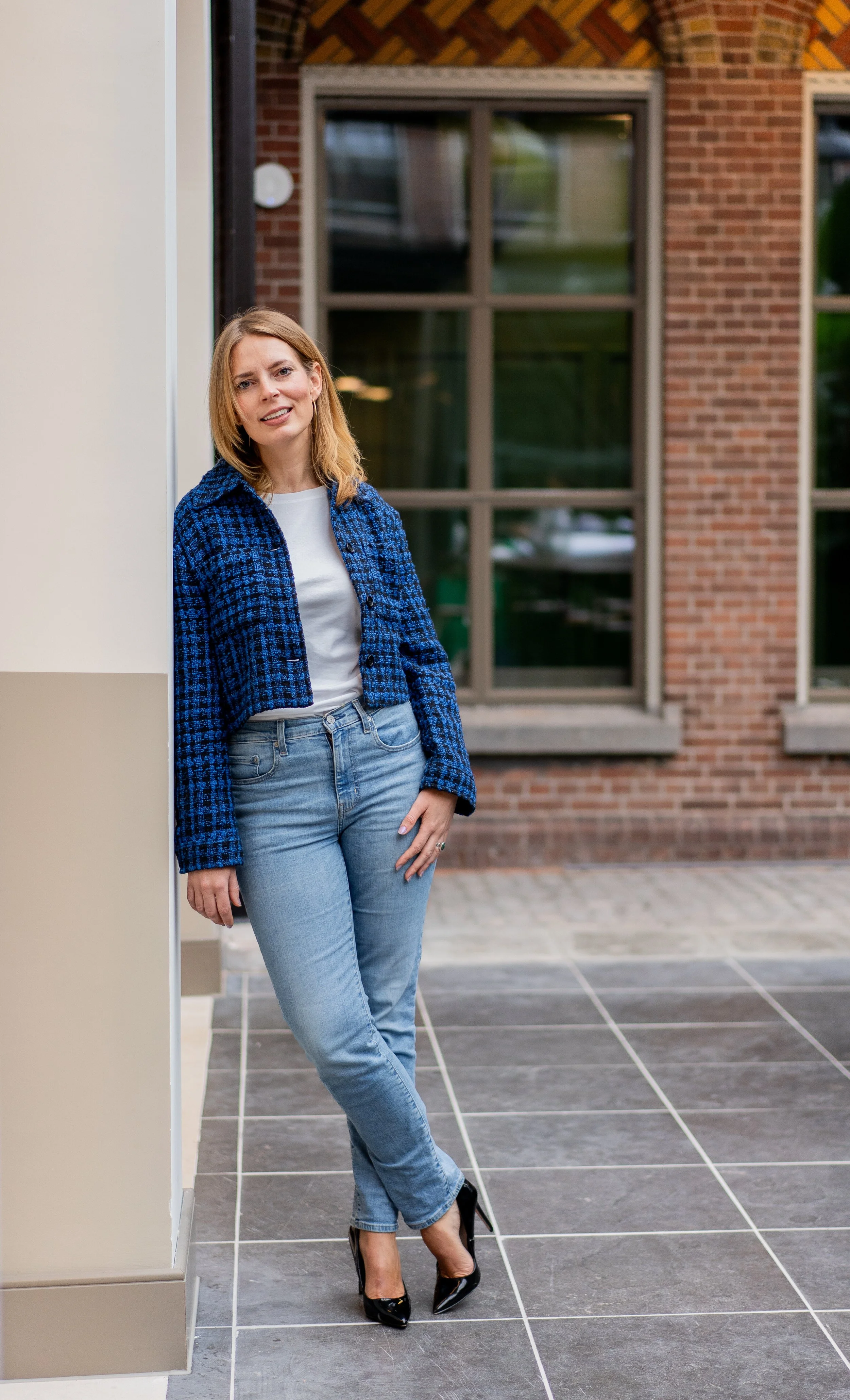 A woman with blonde hair wearing a blue checked jacket, white shirt, light blue jeans, and black high heels standing on a tiled sidewalk outside a brick building with large windows.
