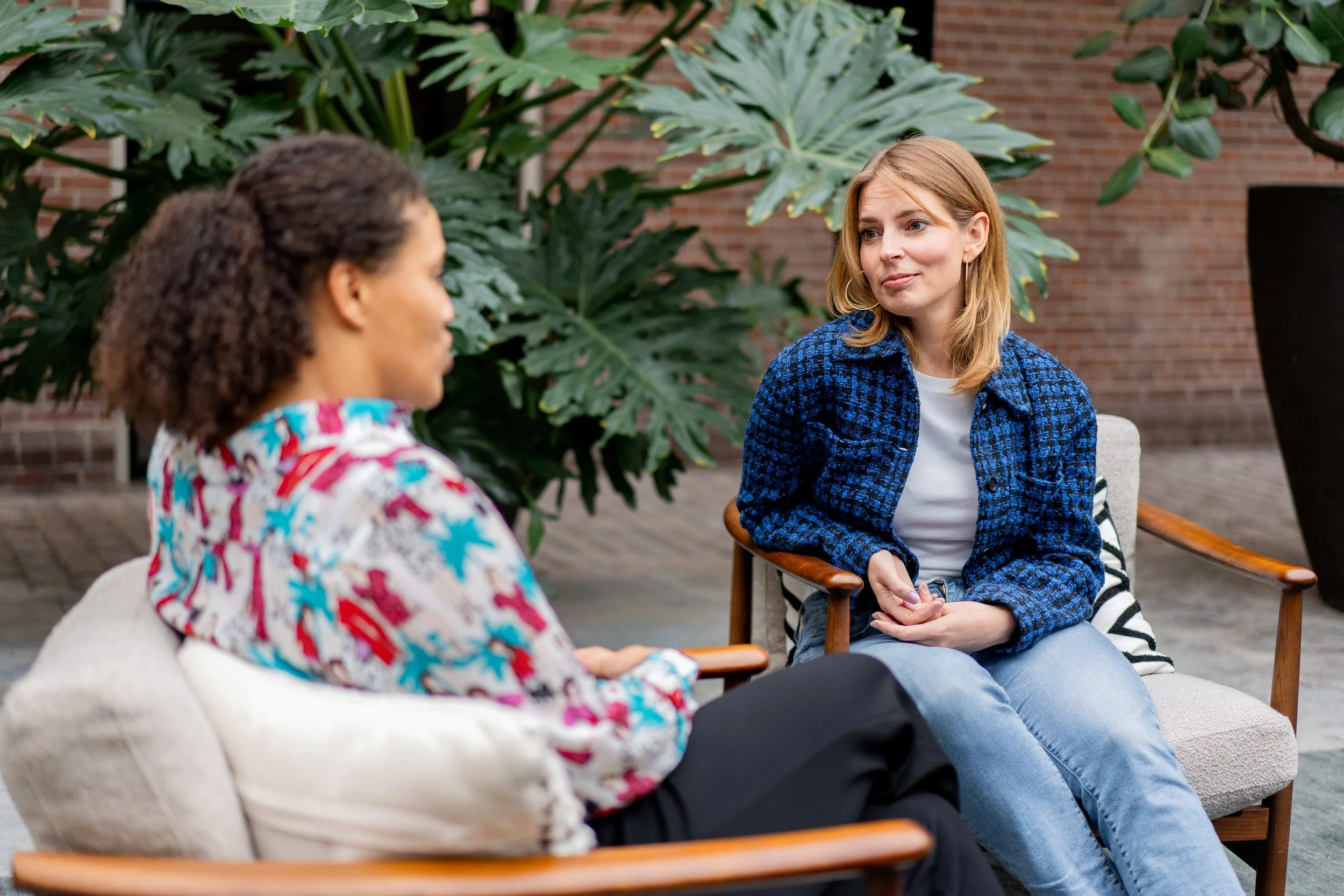 Two women having a conversation in a garden or outdoor space with large leafy plants and a brick wall in the background.