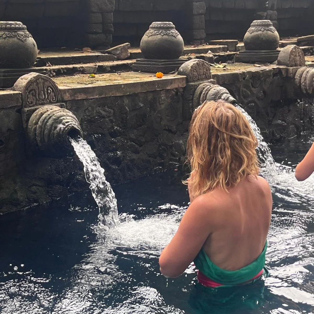 A woman with blonde hair in a green swimsuit standing in a pool near a traditional stone water fountain with water flowing from multiple spouts.