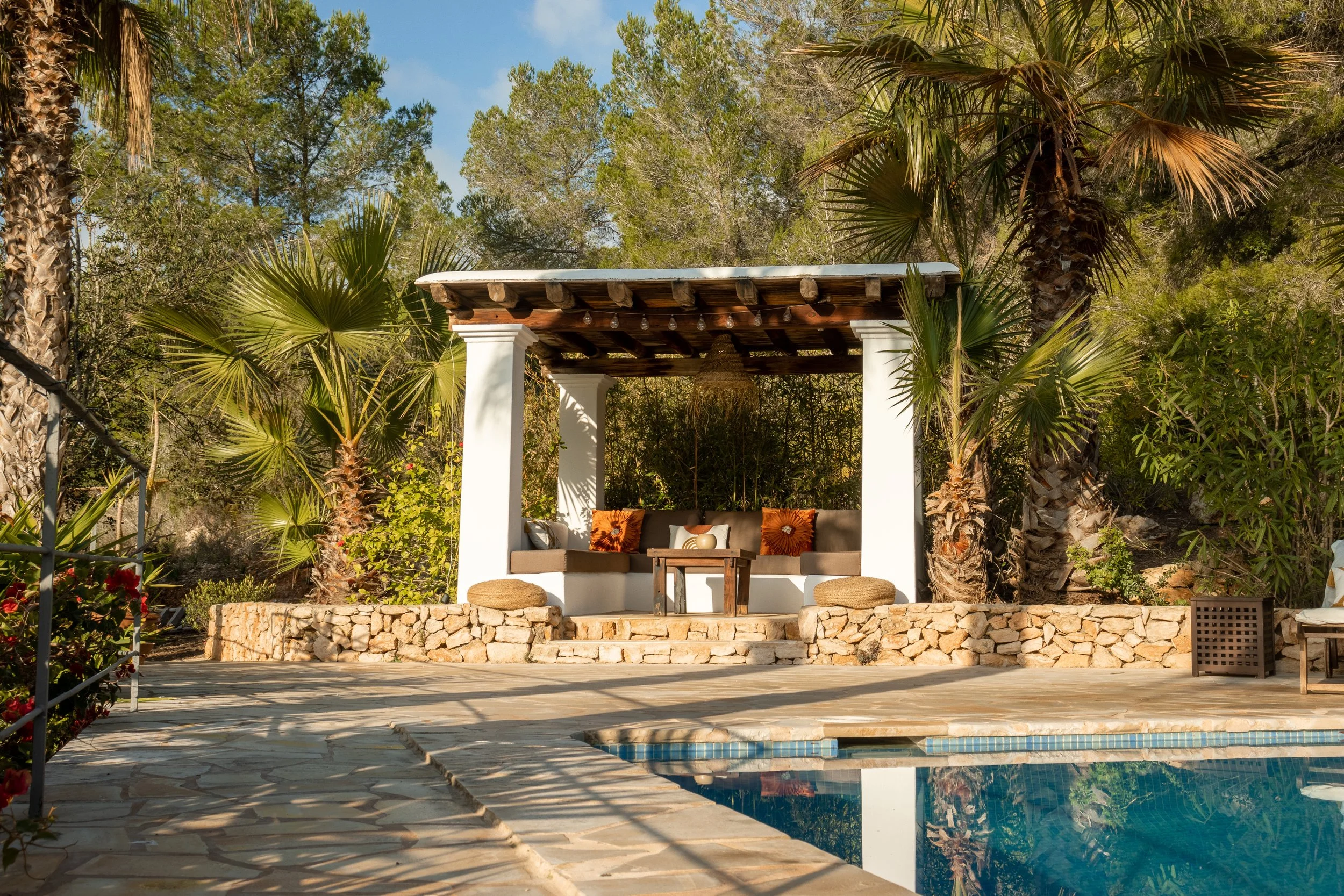 A backyard patio with a swimming pool, a shaded sitting area with cushioned seats, a small table, and surrounding lush greenery including palm trees and shrubs.