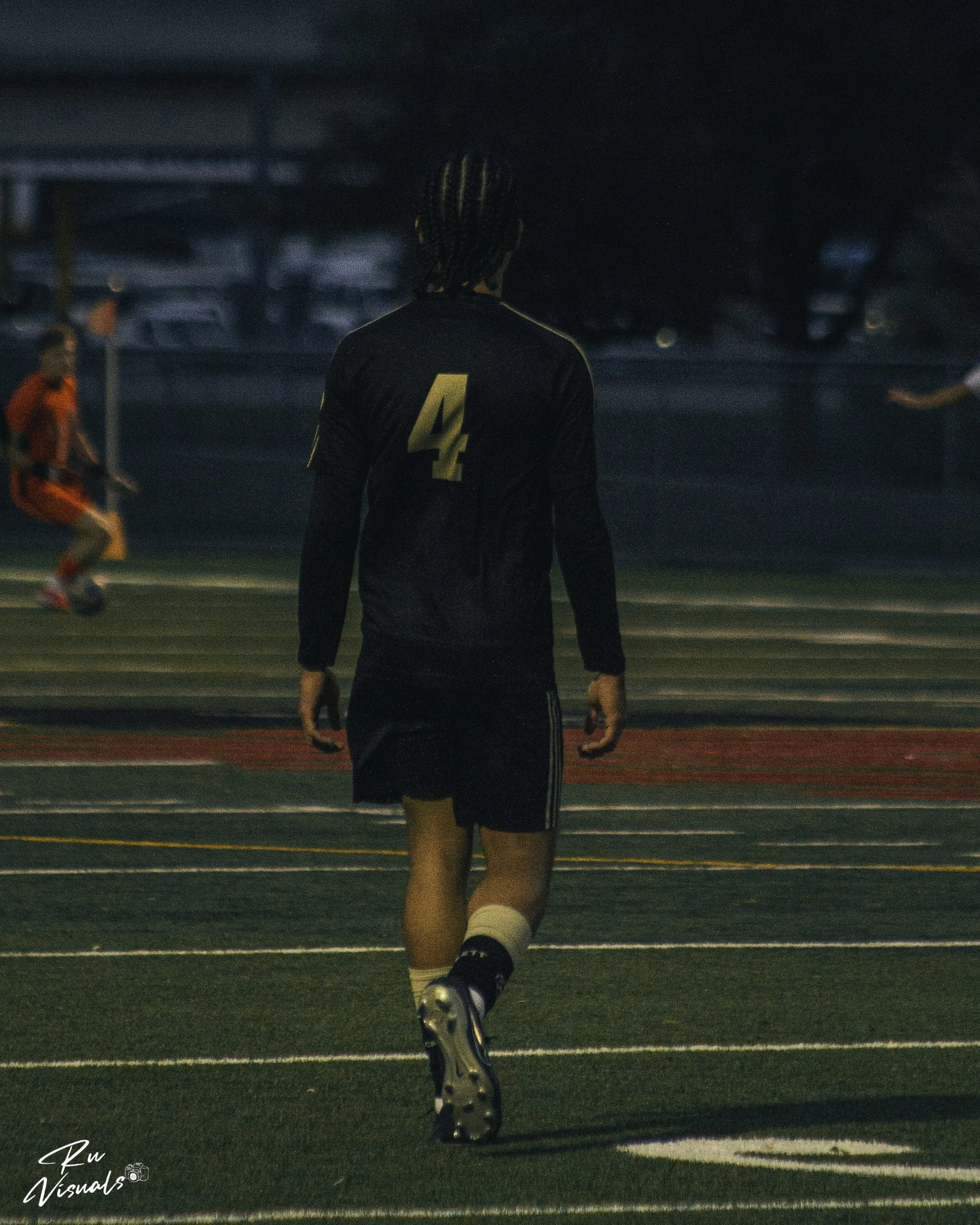A soccer player wearing a black jersey with the number 4 on the back is running on an outdoor field at night. The player has braided hair and is wearing black shorts, white socks, and soccer cleats. There are other players and vehicles in the backgro