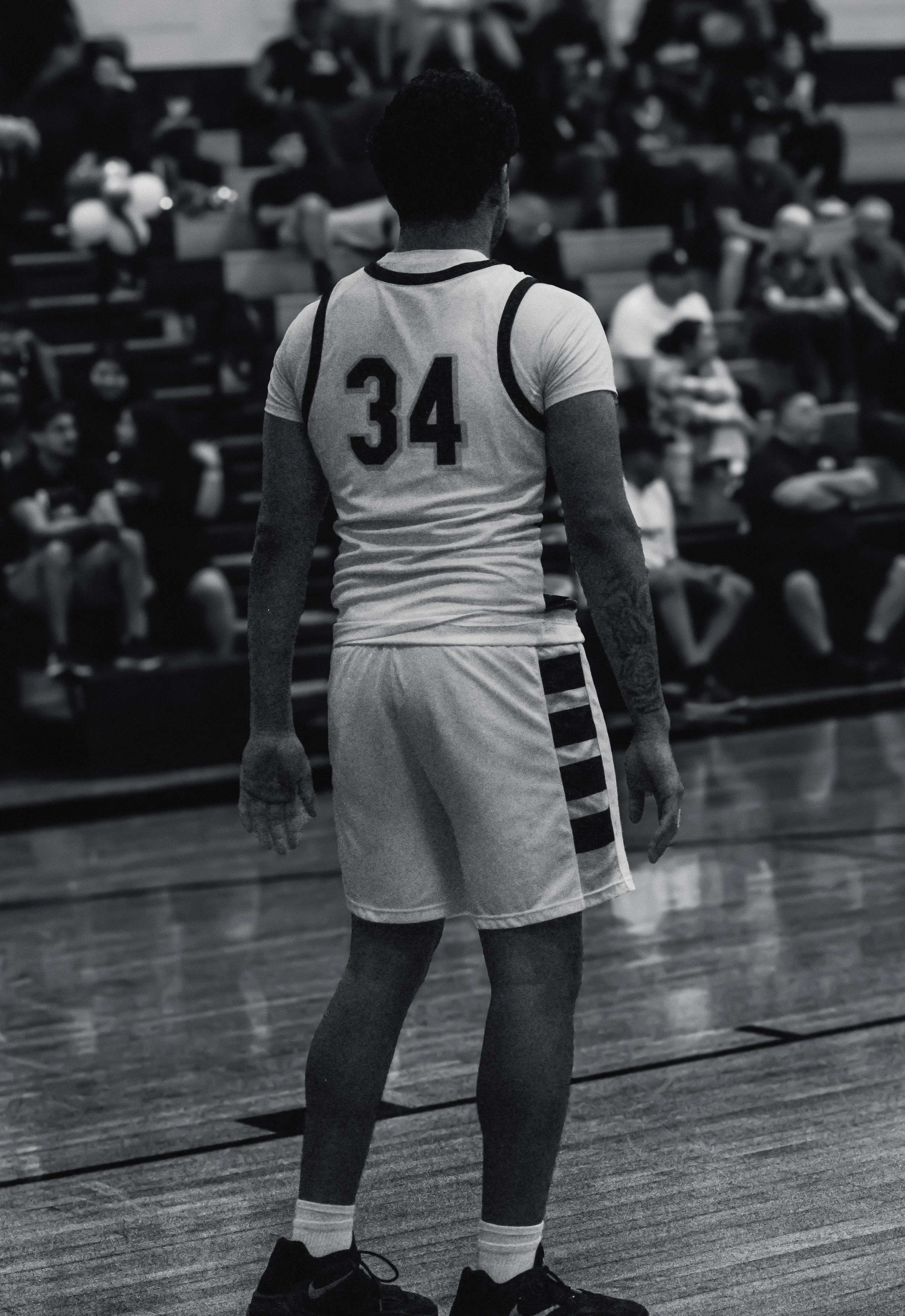 A basketball player wearing a jersey with the number 34 stands on a gym court at a game or practice. The player has short curly hair, and is wearing black Nike shoes, white socks, and shorts. In the background, spectators sit on bleachers.