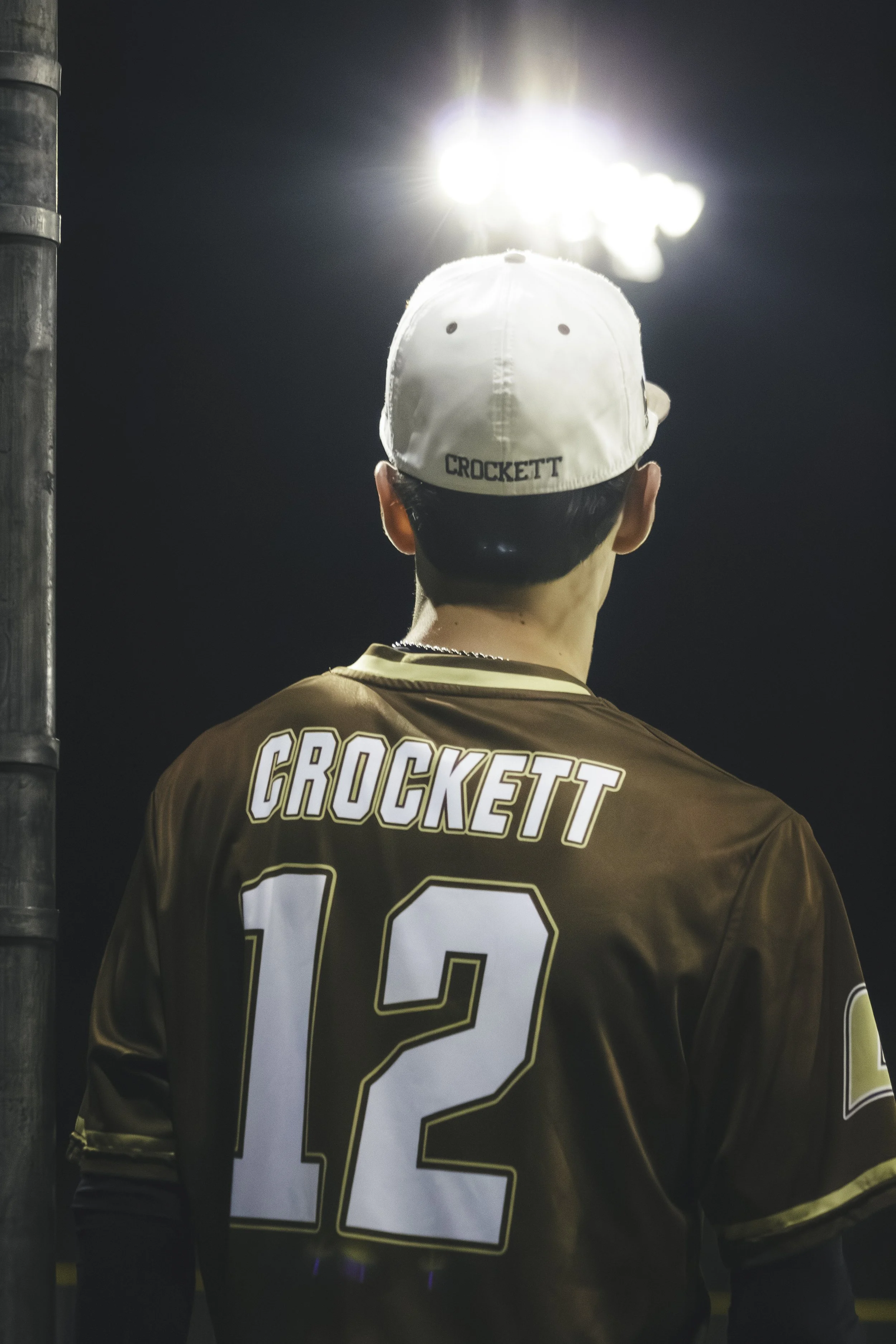 A person wearing a white cap with the name 'CROCKETT' on the back, and a brown sports jersey with the number '13' and the name 'CROCKETT' on it, standing outdoors at night under bright stadium lights.