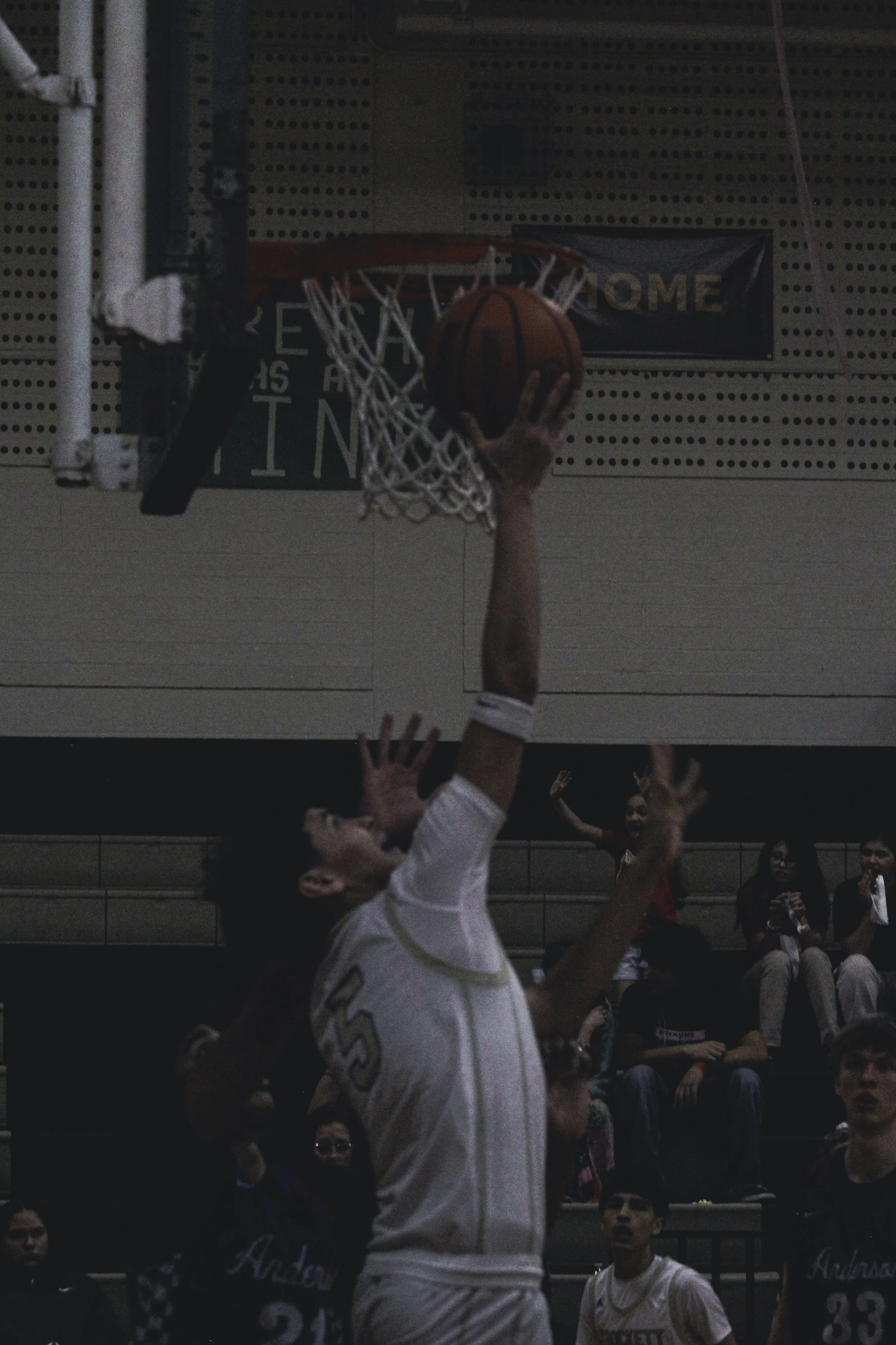 A basketball player in a white uniform is shooting the basketball towards the hoop during a game. Several spectators are sitting and watching in the background.