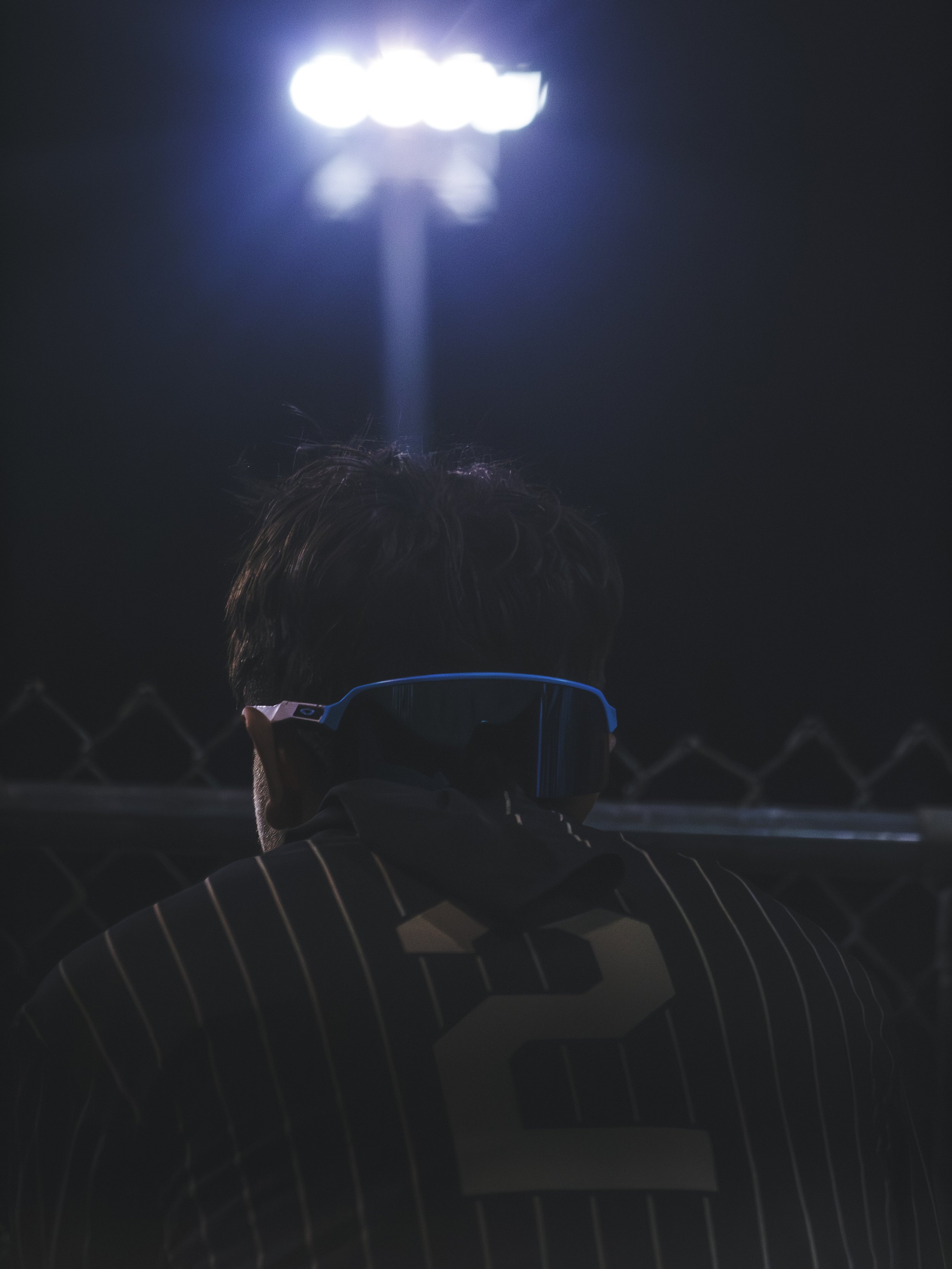 Person with striped shirt wearing glasses, standing by a chain-link fence at night, illuminated by a bright overhead light.