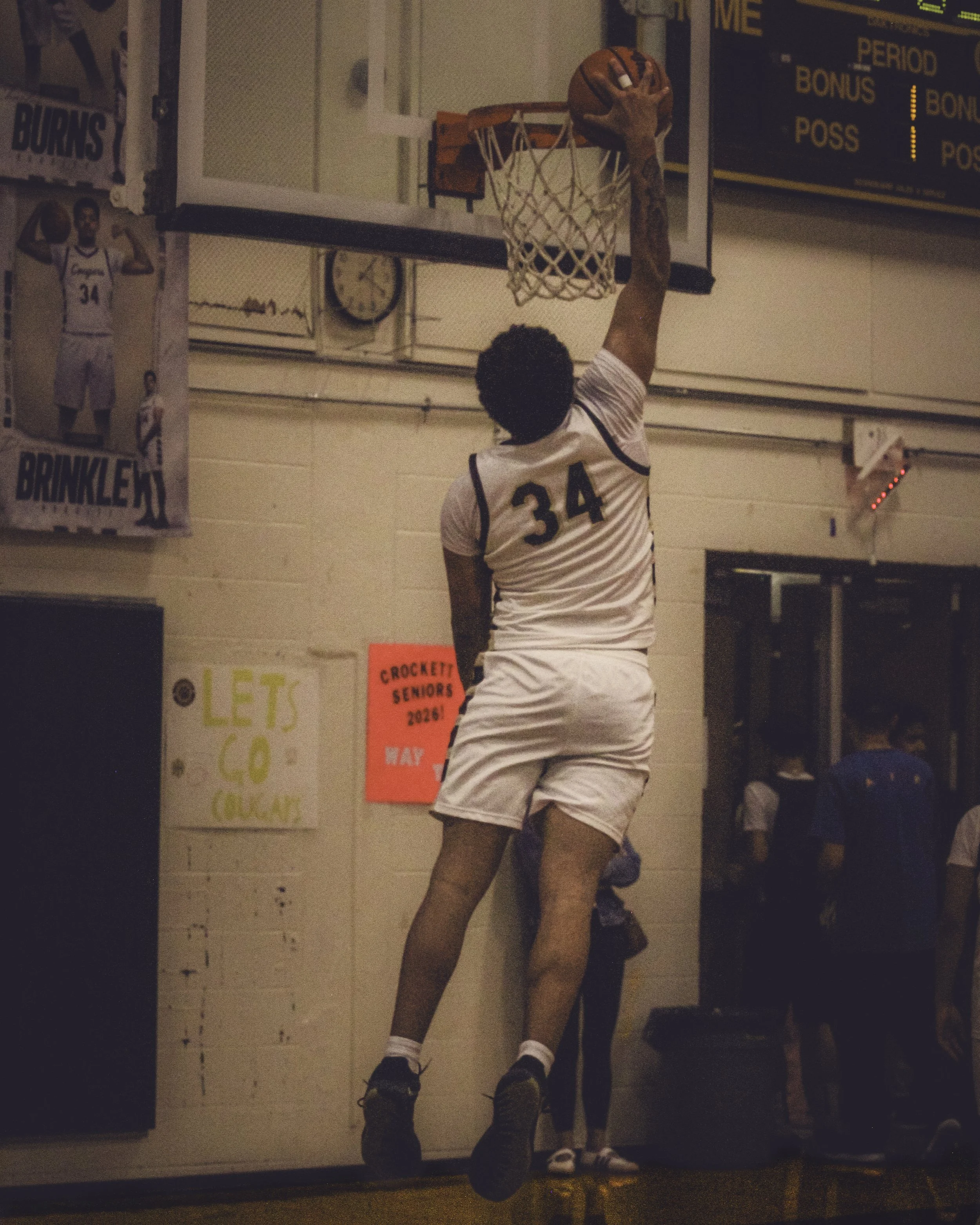 A basketball player wearing a white uniform with the number 34 is dunking a basketball into the hoop. The scene appears to be in an indoor gymnasium with posters and a scoreboard in the background.