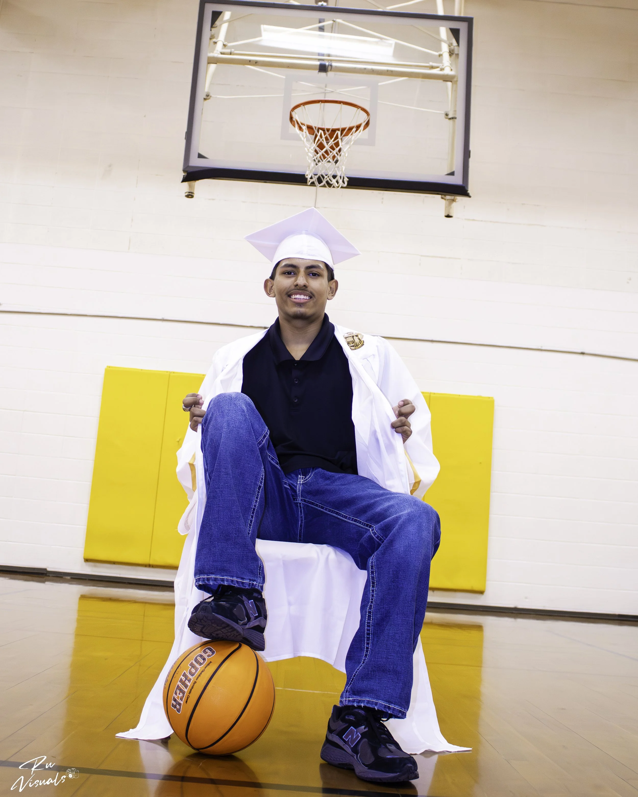 Young man with a graduation cap and gown sitting on a bench in a gymnasium with a basketball in front of him, a basketball hoop hanging above him, and yellow padding on the gym wall behind him.