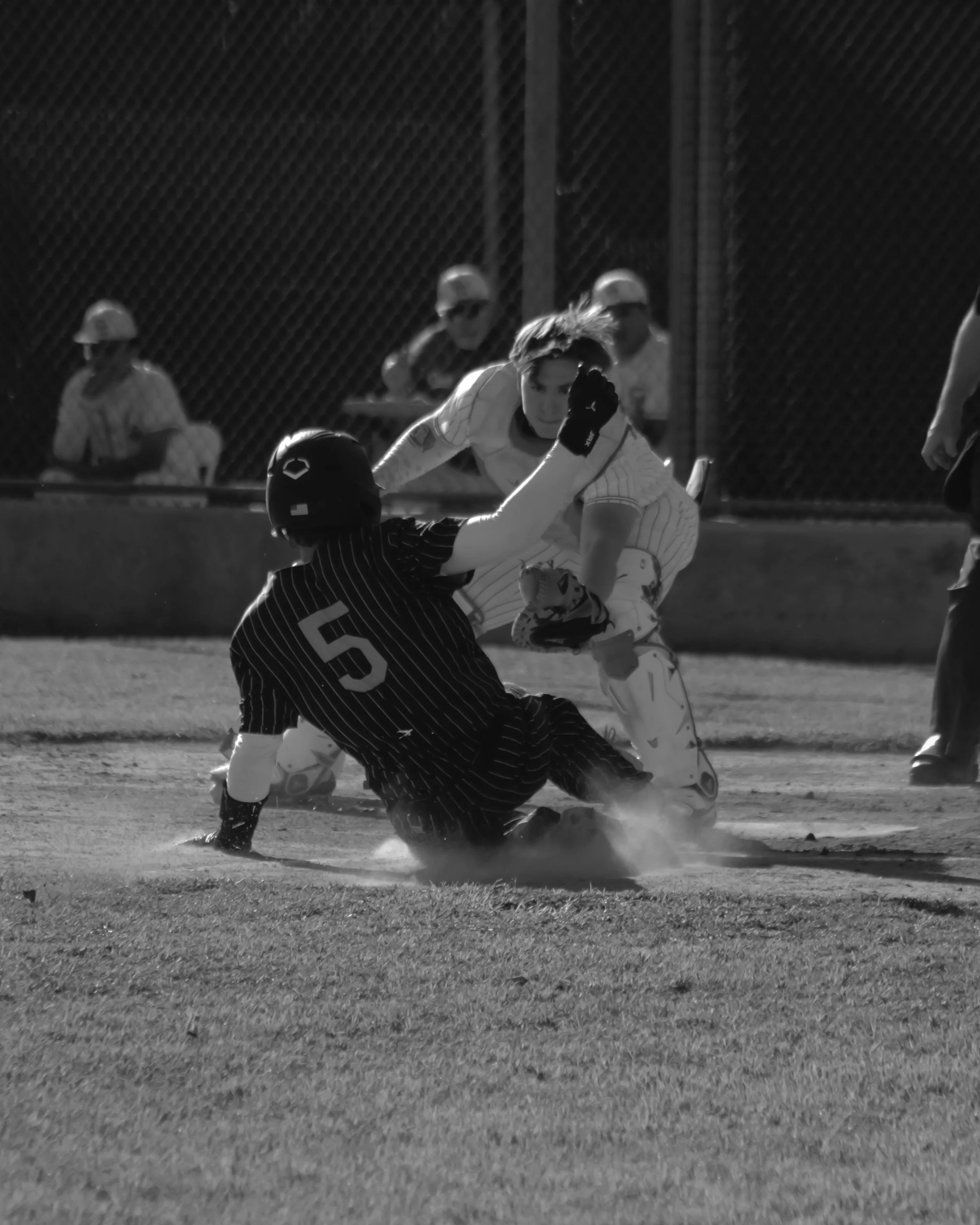 A baseball player sliding into home plate while an opposing catcher attempts to tag him out during a game on a dirt field at night. Several individuals are visible in the background, including two wearing helmets and uniforms, and a chain-link fence 