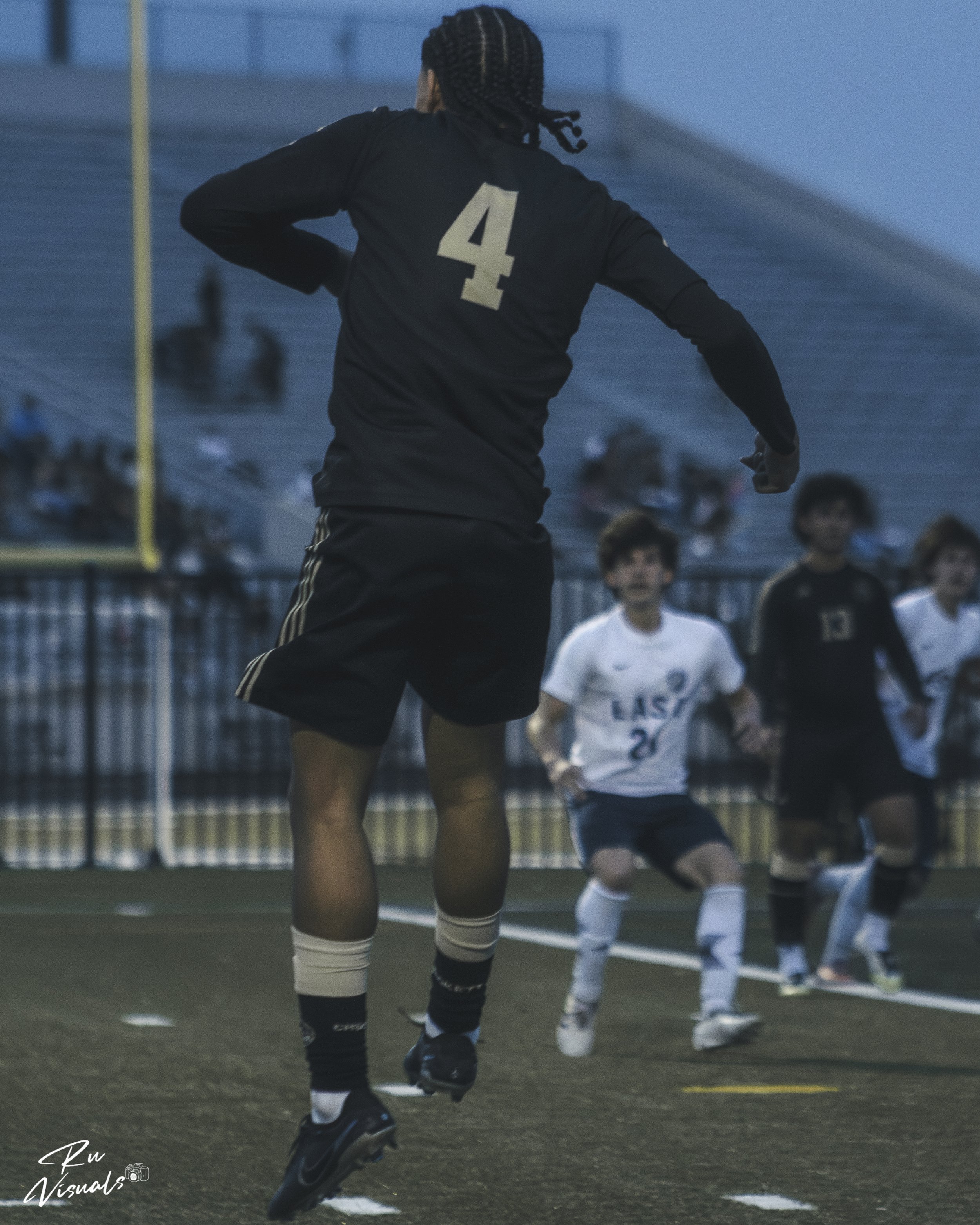 A soccer player in black uniform with the number 4 on the back, running on the field during a game. Two children in white and black uniforms are in the background, watching or waiting for play. The scene appears to be indoors with a blue wall and met