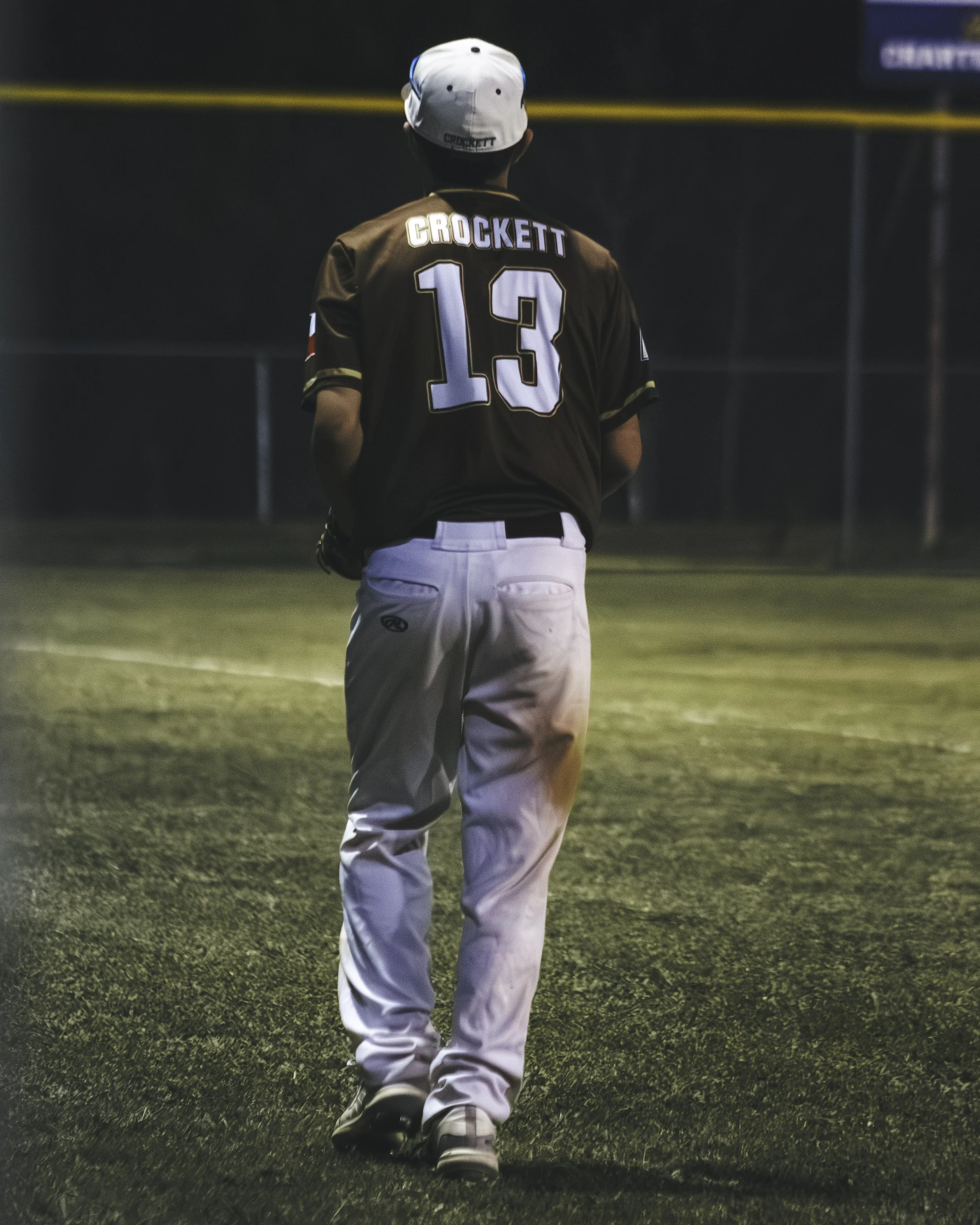A baseball player walking on the field wearing a jersey with the name 'CROCKETT' and the number '13' on the back, white pants, a white cap, and a black glove on his left hand.