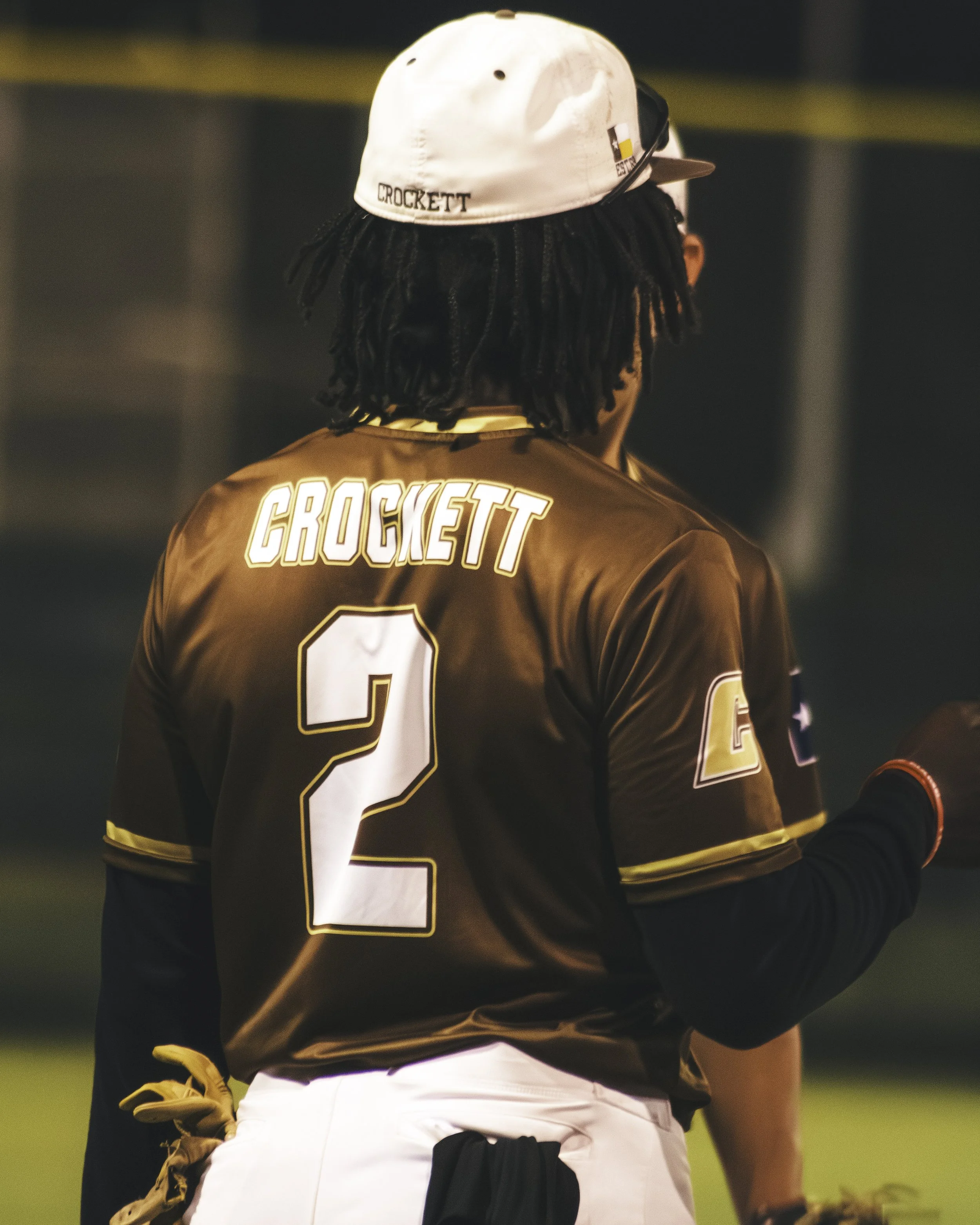 A person wearing a white baseball cap with the name 'CROCKETT' on the back, a black sports jersey with 'CROCKETT' and the number 2, and white pants with black and yellow accents, appears to be on a sports field at night.