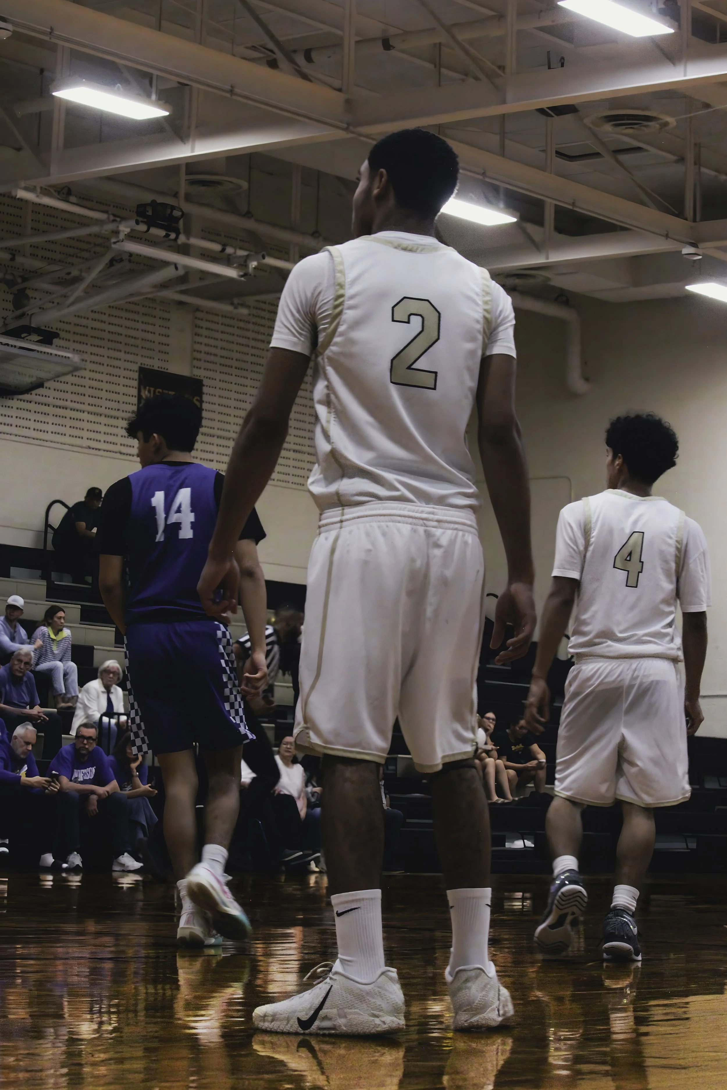 Basketball players in white and blue uniforms on a wooden court, with spectators seated in the background.