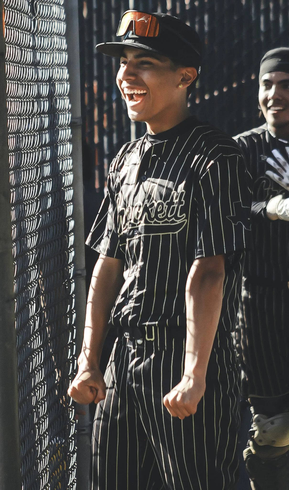 A baseball player in a black and white pinstriped uniform smiling, wearing a black cap and sunglasses on top, standing next to a chain-link fence.
