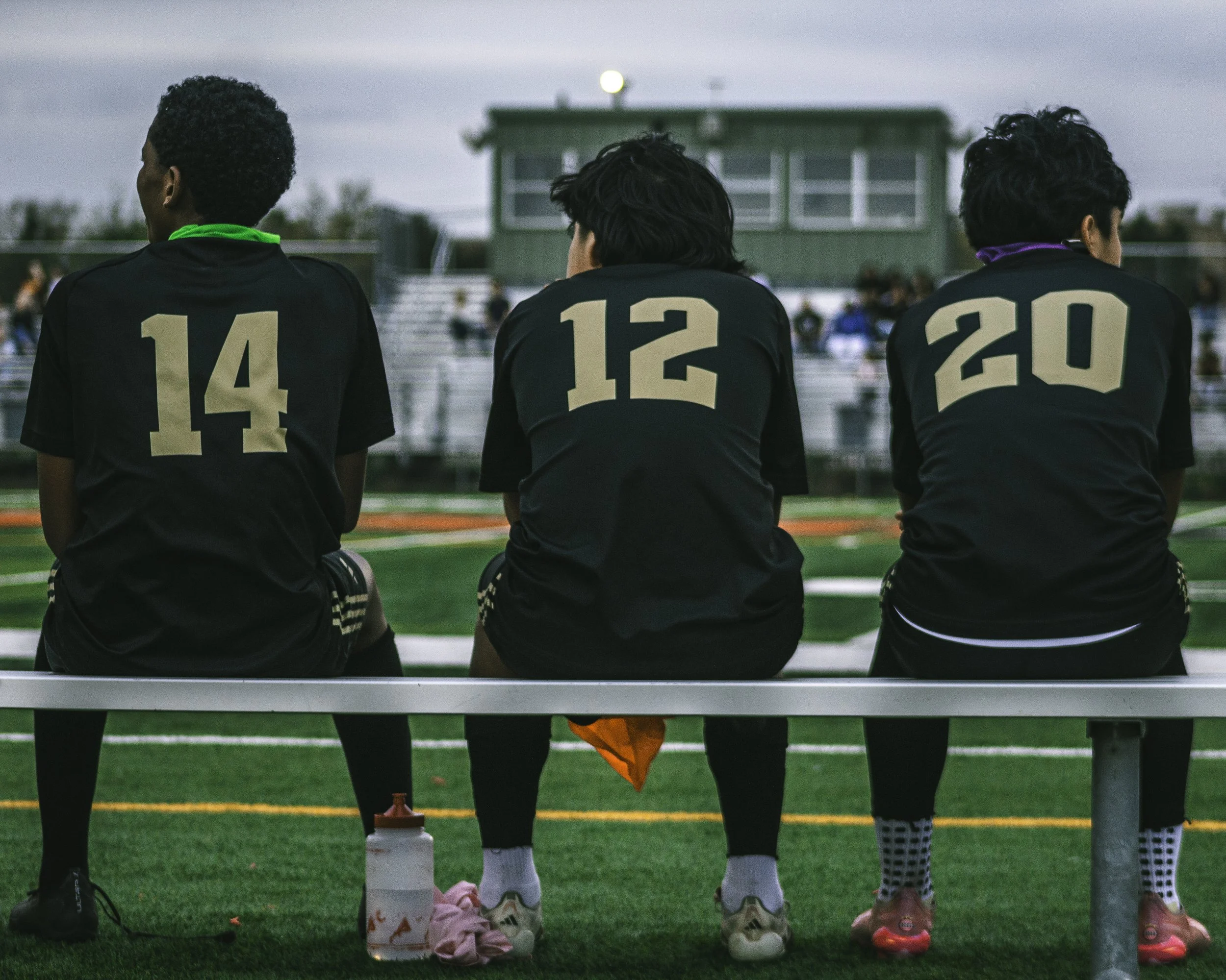 Three soccer players with numbers 14, 15, and 20 on their black jerseys sitting on a bench on a soccer field, viewed from behind.