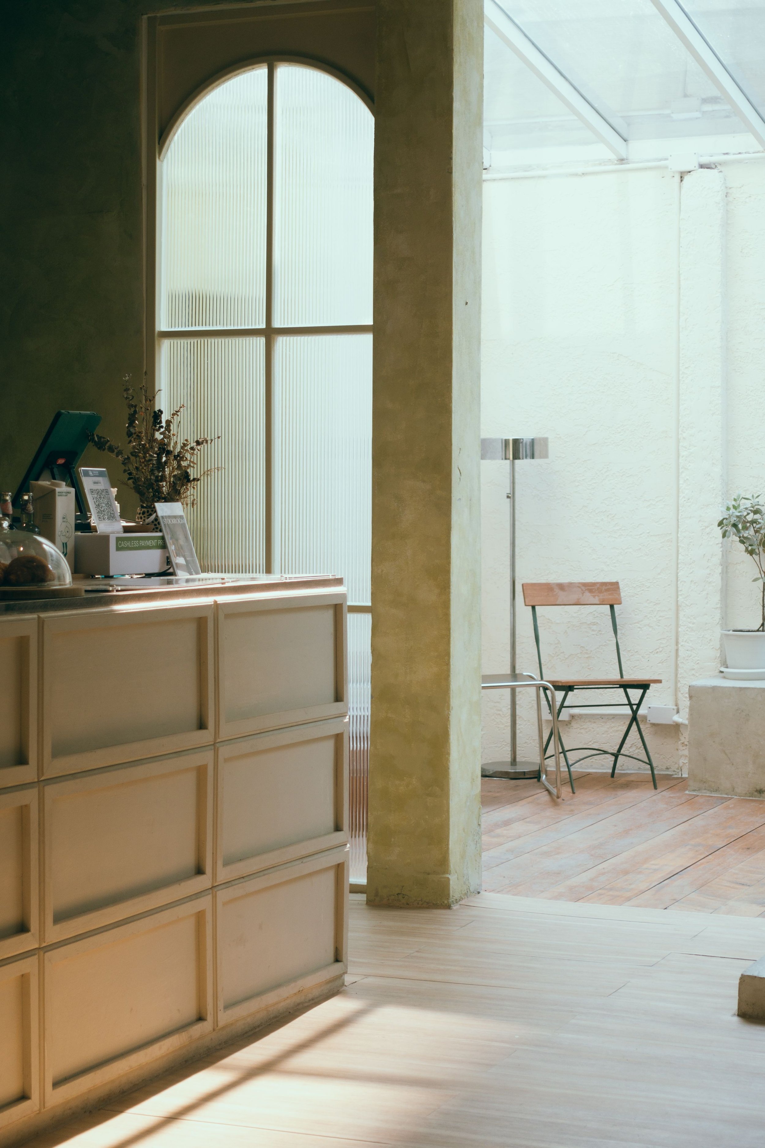 Interior of a room with a reception counter on the left, a large frosted window/door with an arched top behind it, and a sitting area with two chairs and a lamp on the right. There are some books and decorations on the counter, and potted plants near the window.