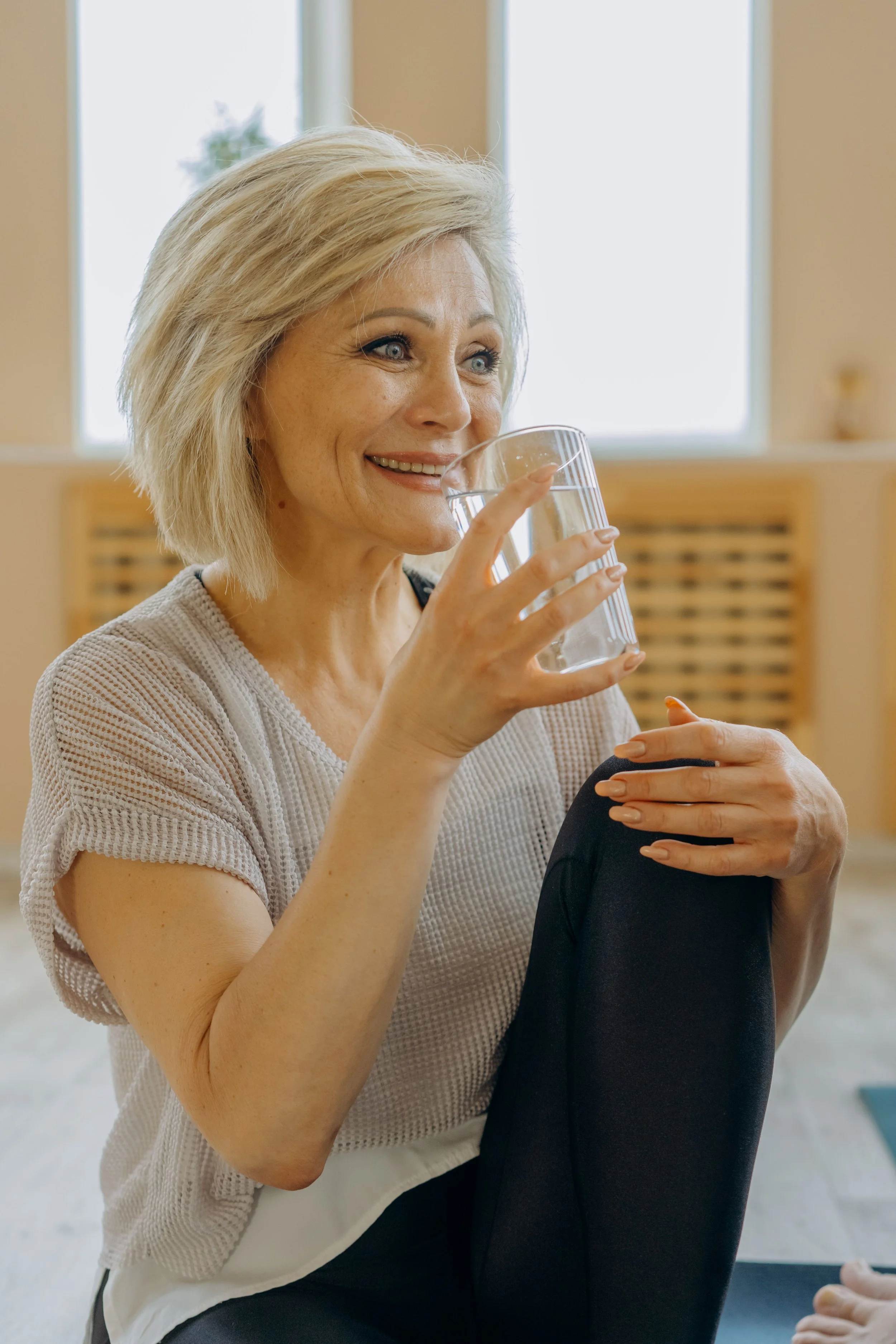 A woman with blonde hair smiling and drinking water from a clear glass while sitting with a person in a yoga pose with their legs in a butterfly position.